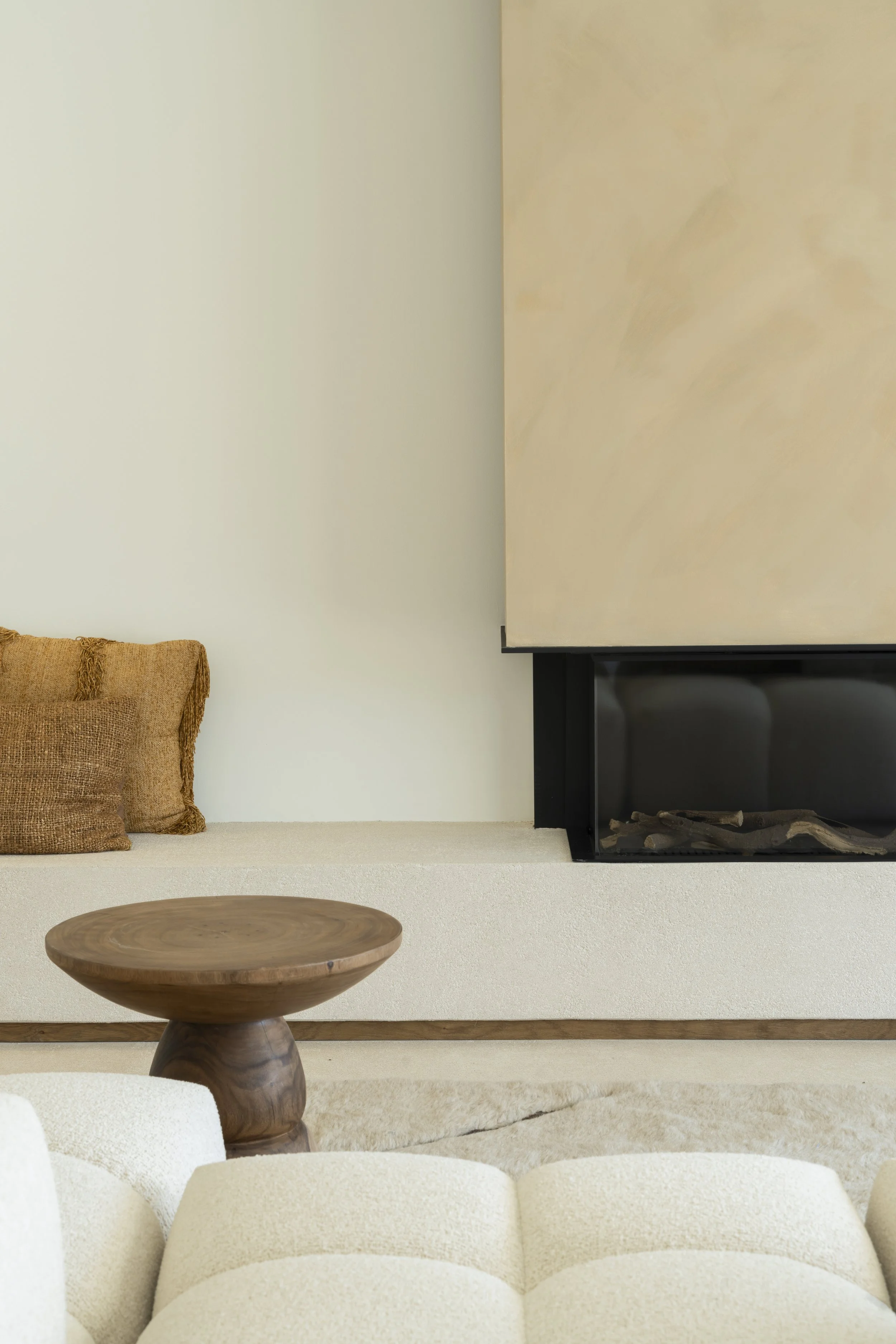 Living room thoughtfully picked out items from our interior store at the costa blanca, Moraira. Beige sofa, wooden round coffee table, and a textured cushion, against a white wall with fireplace.