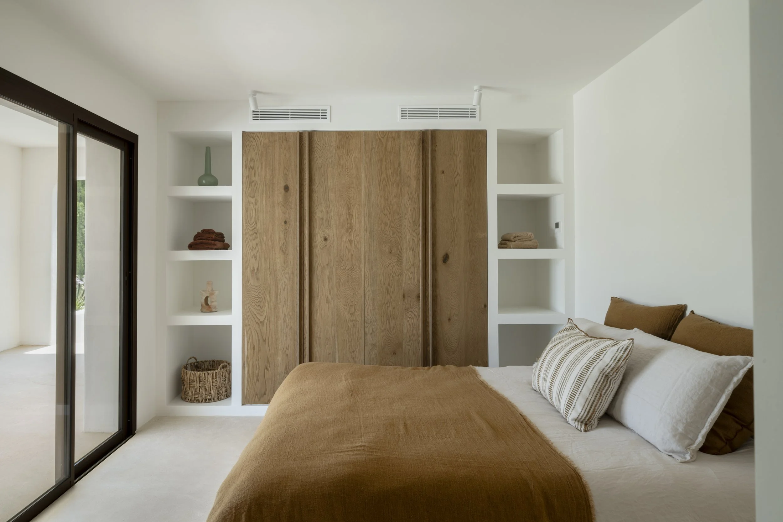 Modern bedroom with a bed dressed in white linens and brown accents, a built-in wooden closet, and open shelves with decorative items, adjacent to a sliding glass door leading to an outdoor space.