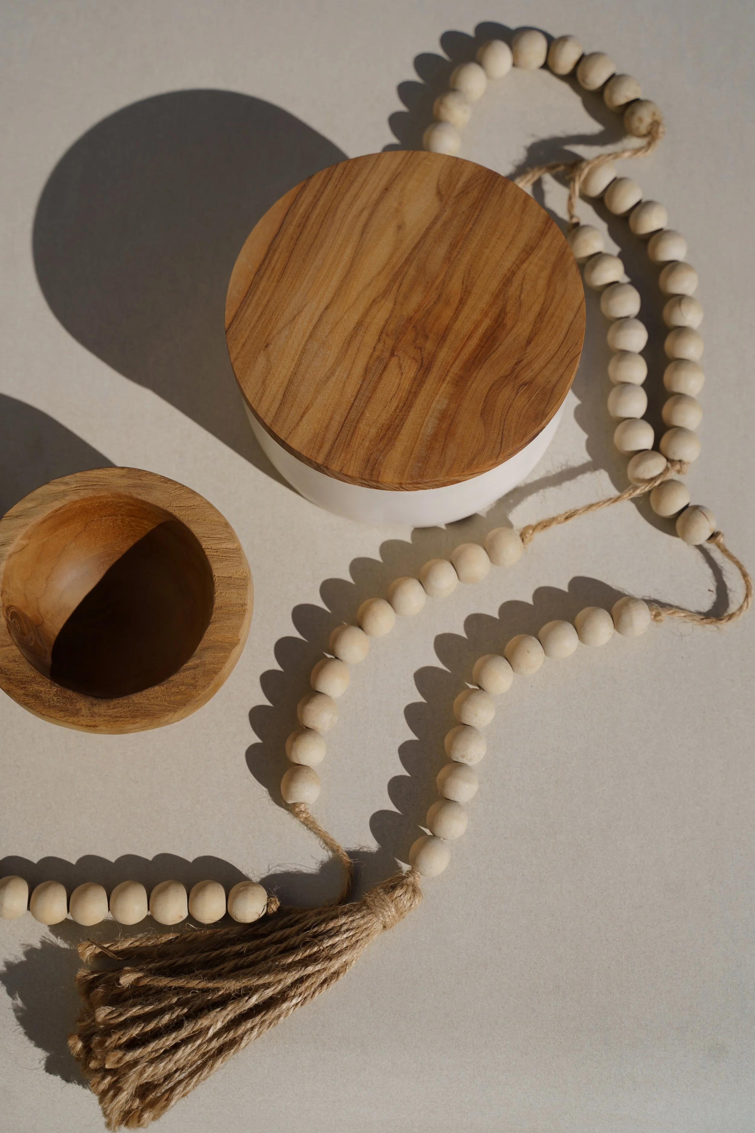 You can find these items in our store A Concept store,  round wooden lid, a small wooden bowl, and a beaded rope with a tassel lie on a flat surface, casting shadows.