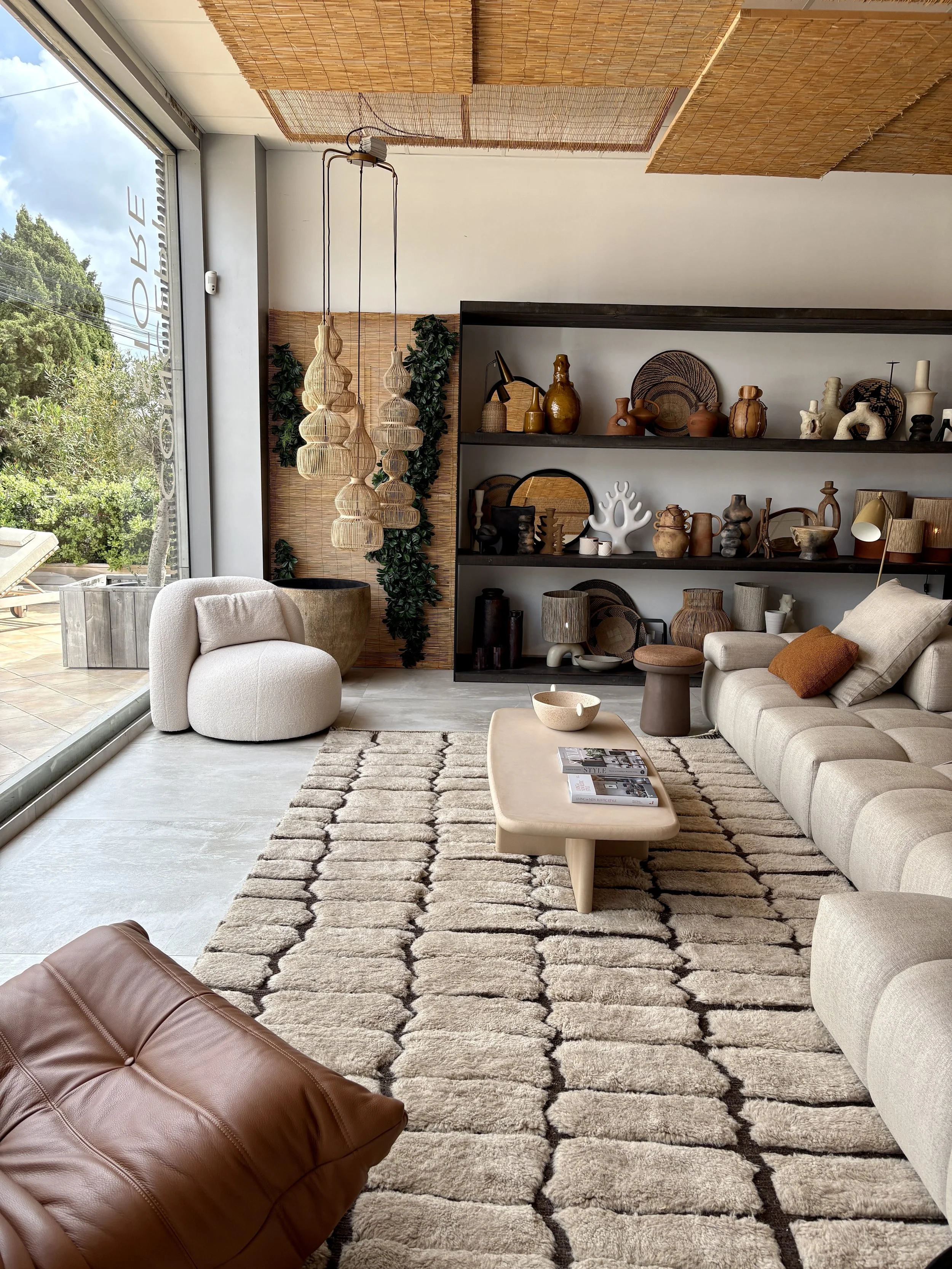 Modern living room with beige sectional sofa, plush cream armchair, brown leather ottoman, textured rug, and floating shelves displaying pottery and decorative items. Large glass window with outdoor greenery.