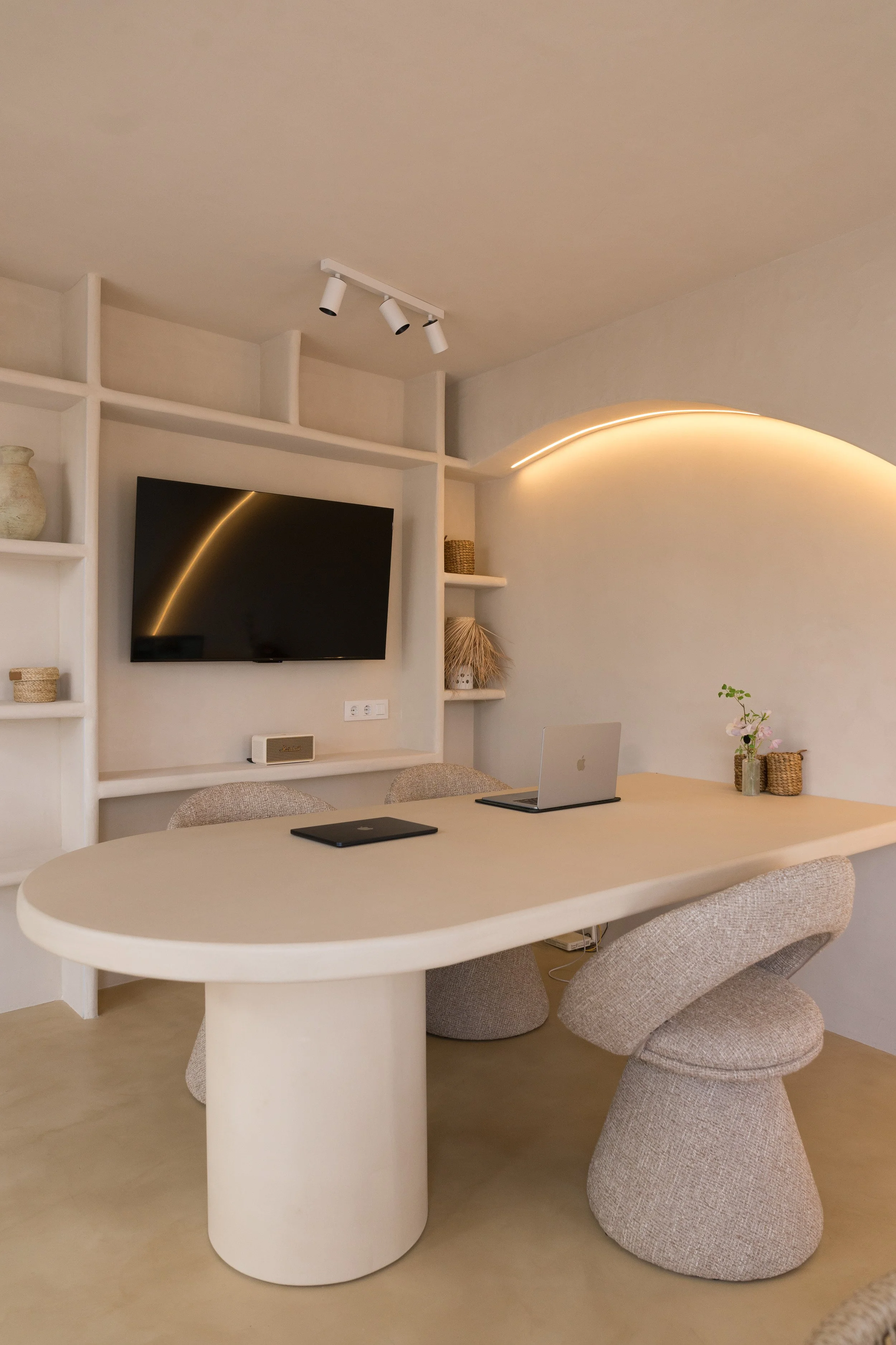 Modern dining room with oval table, four curved beige chairs, wall-mounted TV, built-in shelves with decorative items, and minimalist lighting.
