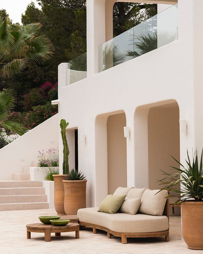 Outdoor patio with a beige sofa with pillows, potted plants including a tall cactus, decorative bowls, and a staircase leading to a balcony with a glass railing, set against a white building and green trees.