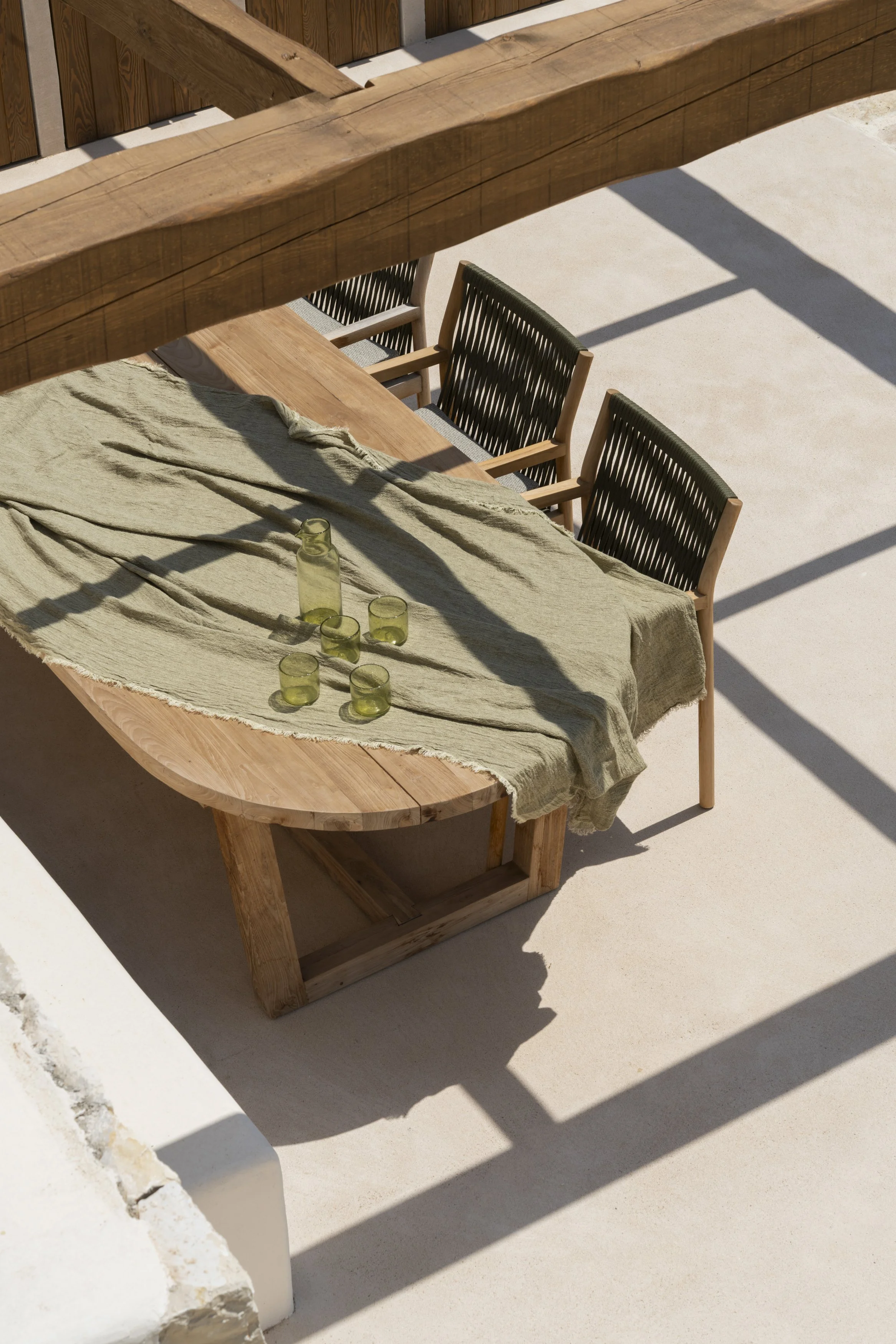 Outdoor dining table with beige tablecloth and four green glasses, shaded by wooden beams, with wooden chairs in the background.