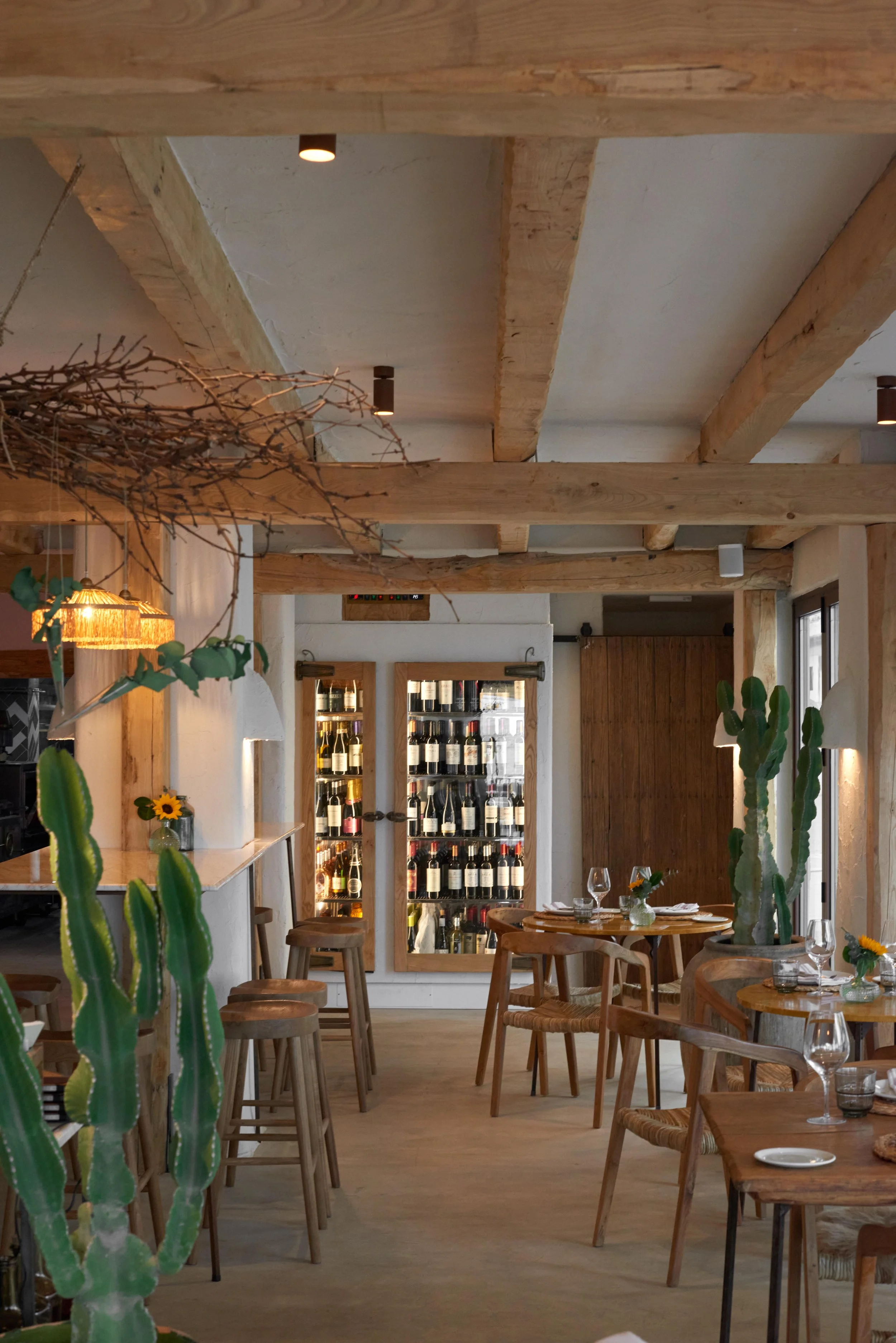 Interior of a cozy restaurant with wooden beams, potted cacti, and a wine display in the background, set for dining.