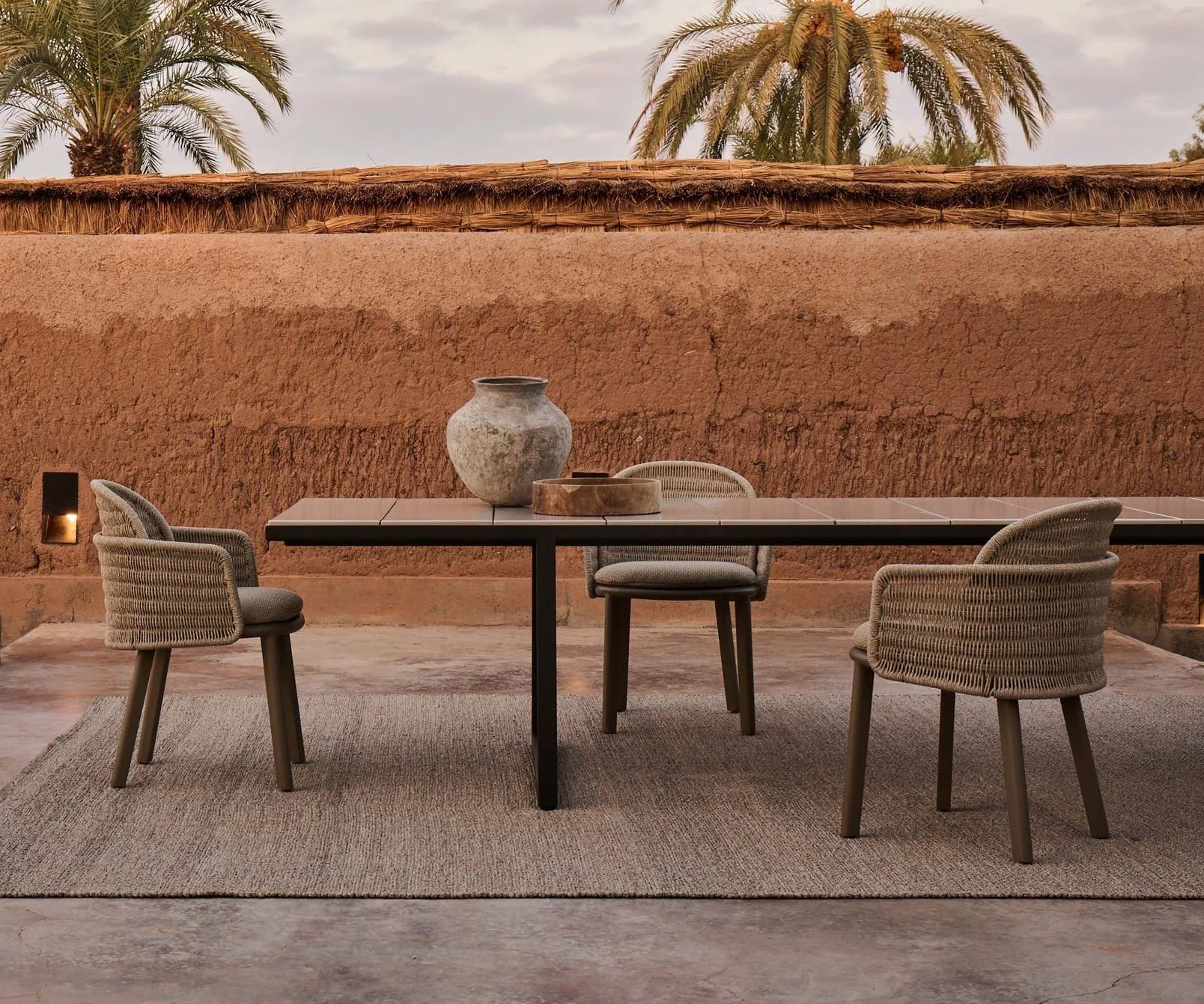 An outdoor patio with a long rectangular table, four woven chairs, a large ceramic vase, and a wooden tray with a smaller bowl. The background features a textured mud wall, palm trees, and a cloudy sky.