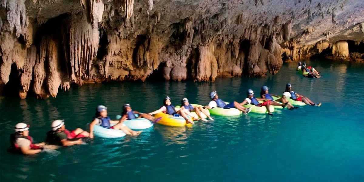 Group of people in life jackets on tubes floating in a cave with stalactites hanging from the ceiling and calm water.