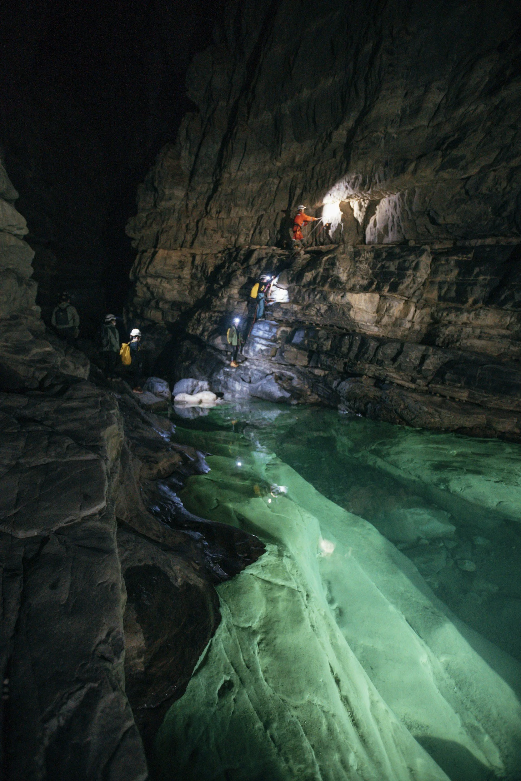 A group of people exploring a dark, narrow cave with a water-filled floor, some individuals climbing on rocks while others are observing, illuminated by flashlights and headlamps.