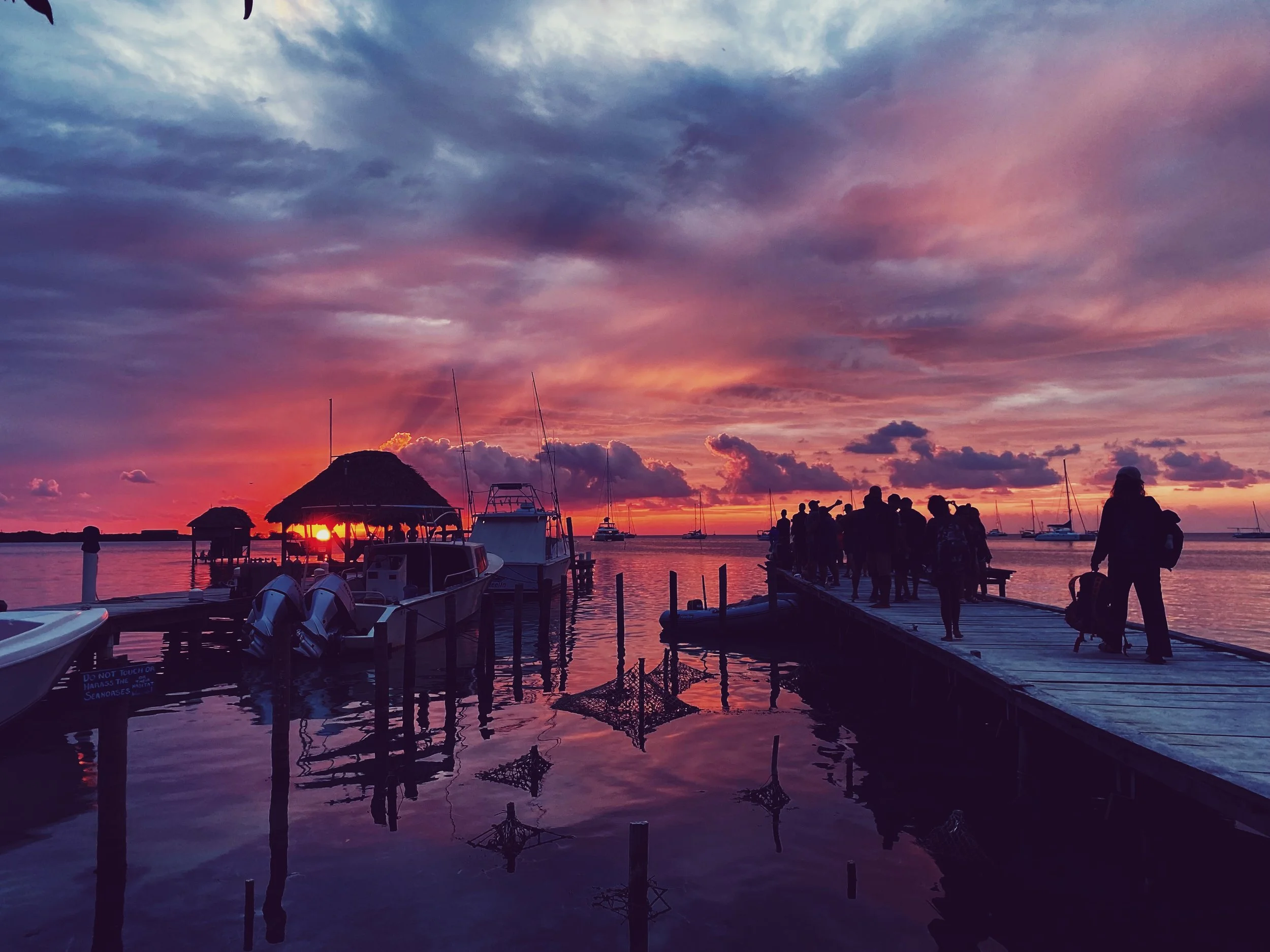 Silhouettes of people walking on a dock during sunset with boats and a thatched-roof structure in the water