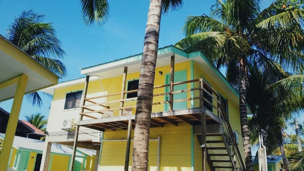 Yellow house with a balcony under construction, surrounded by tall palm trees, against a bright blue sky.