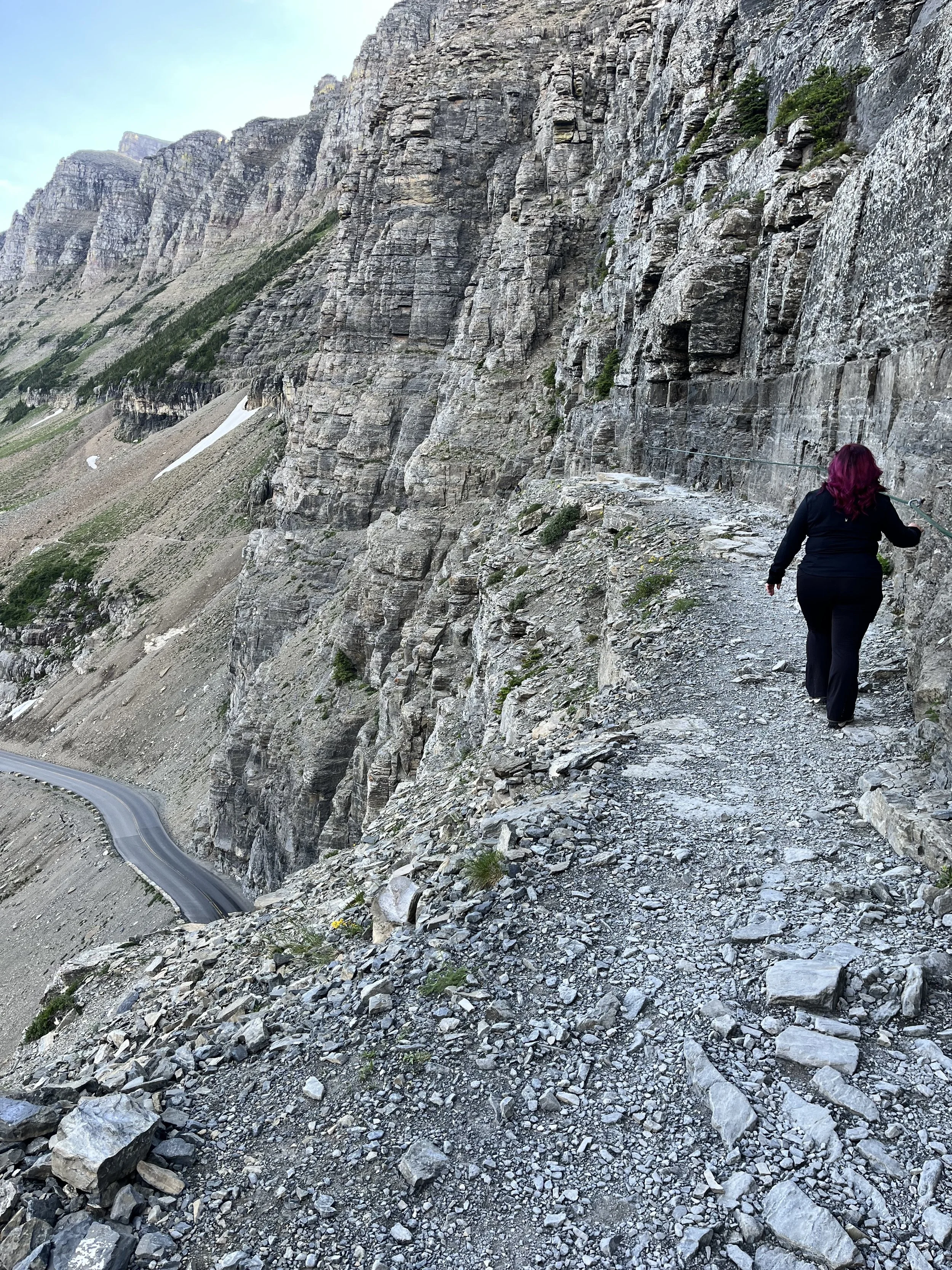 A woman with purple hair wearing black clothing hiking on a narrow rocky trail along a cliffside with a mountain landscape in the background.