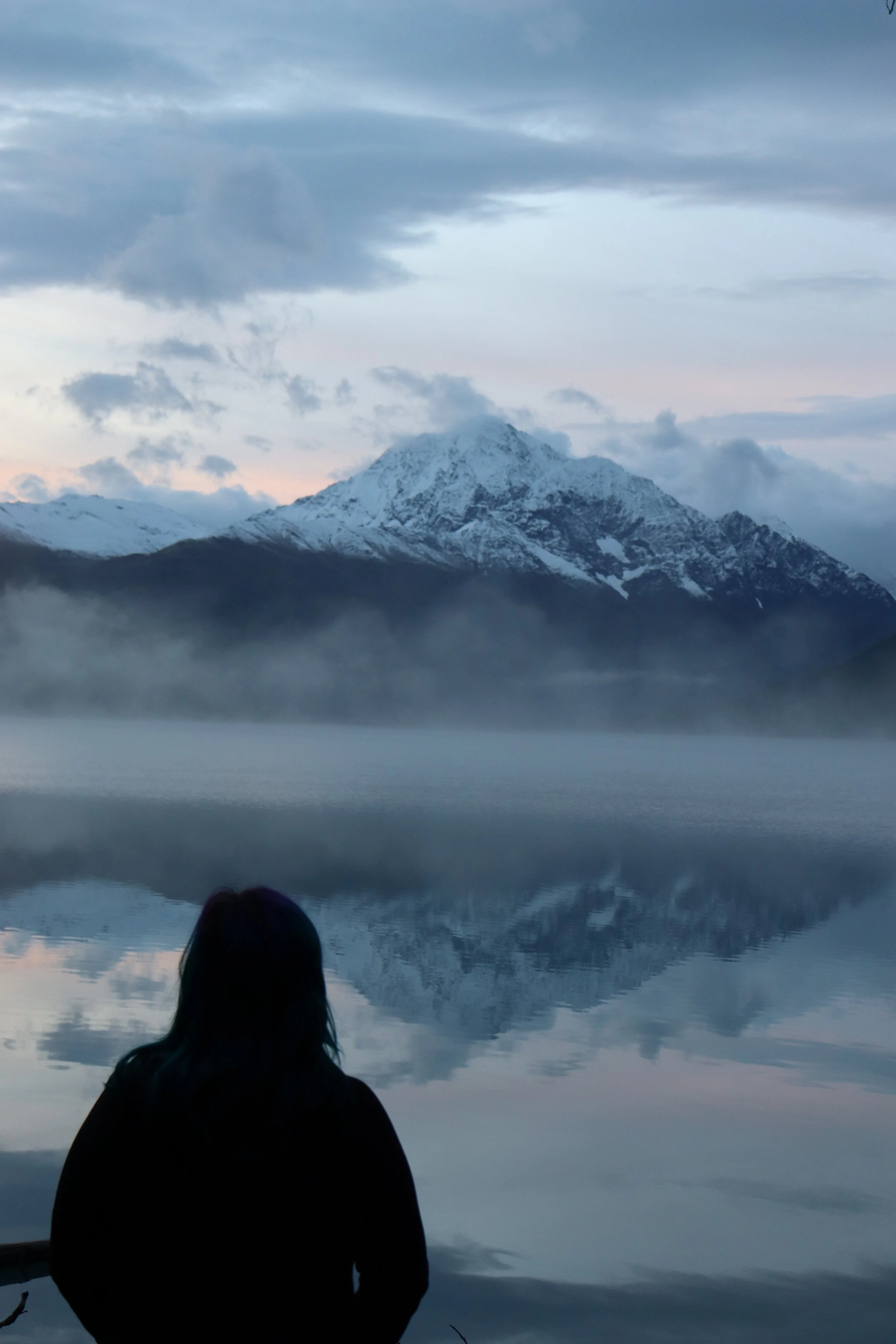 A person sitting by a calm lake, looking at a snow-capped mountain in the distance, with a misty atmosphere and cloudy sky.