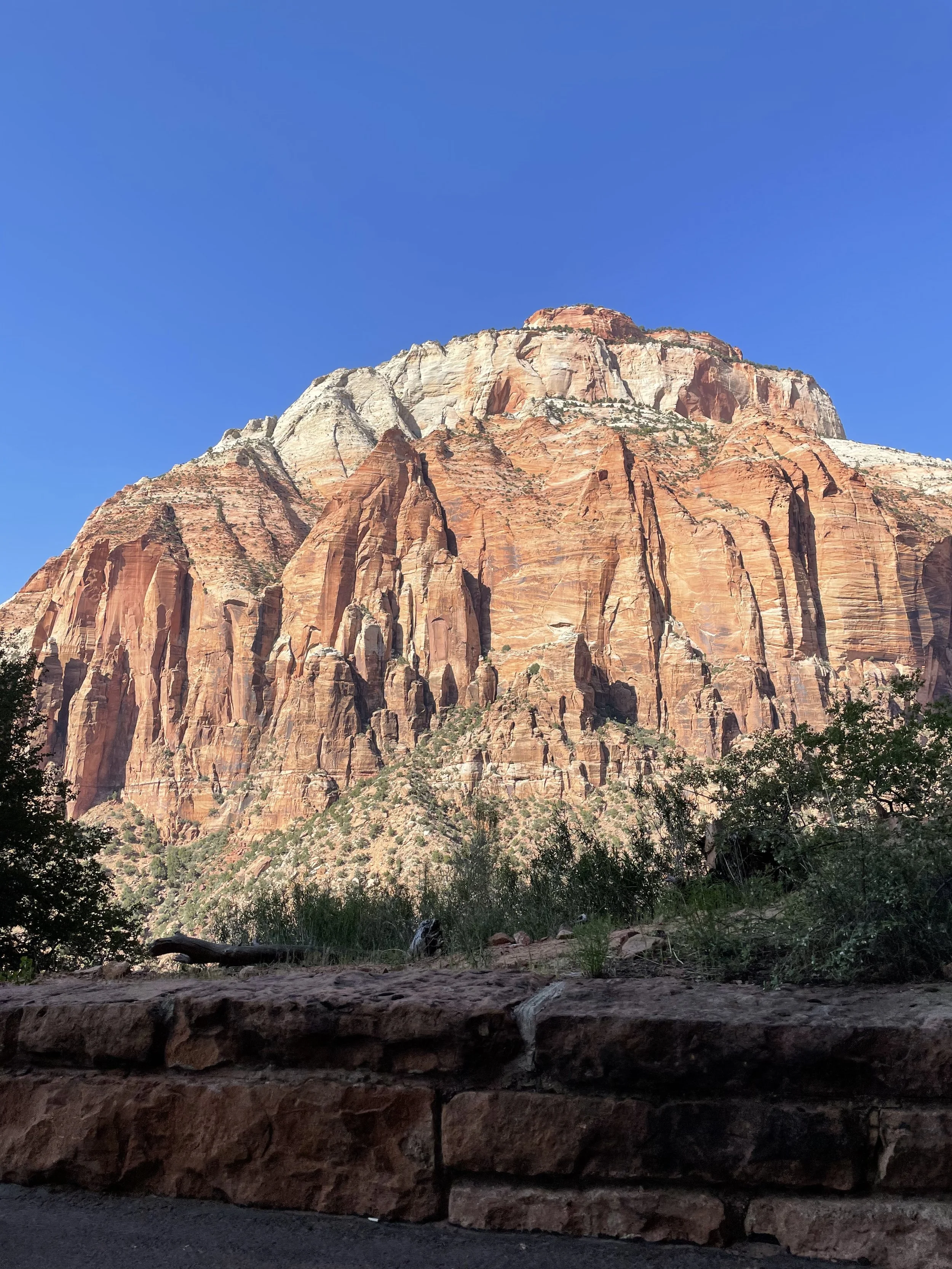 A large red rock mountain under a clear blue sky with some green vegetation and a stone wall in the foreground.