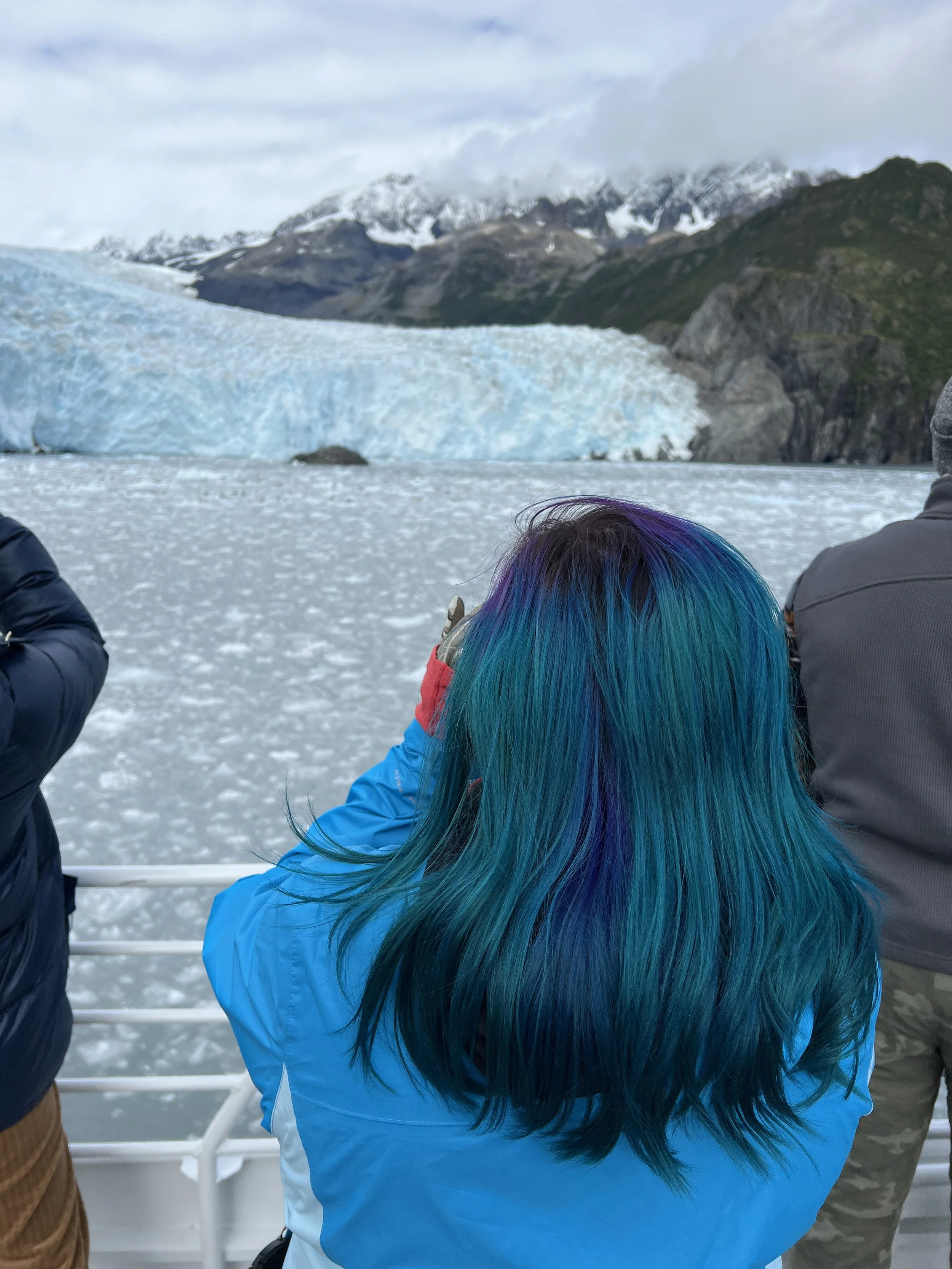 Person with colorful blue, green, and purple hair wearing a blue jacket taking a photo of a glacier and mountains from a boat with other people.