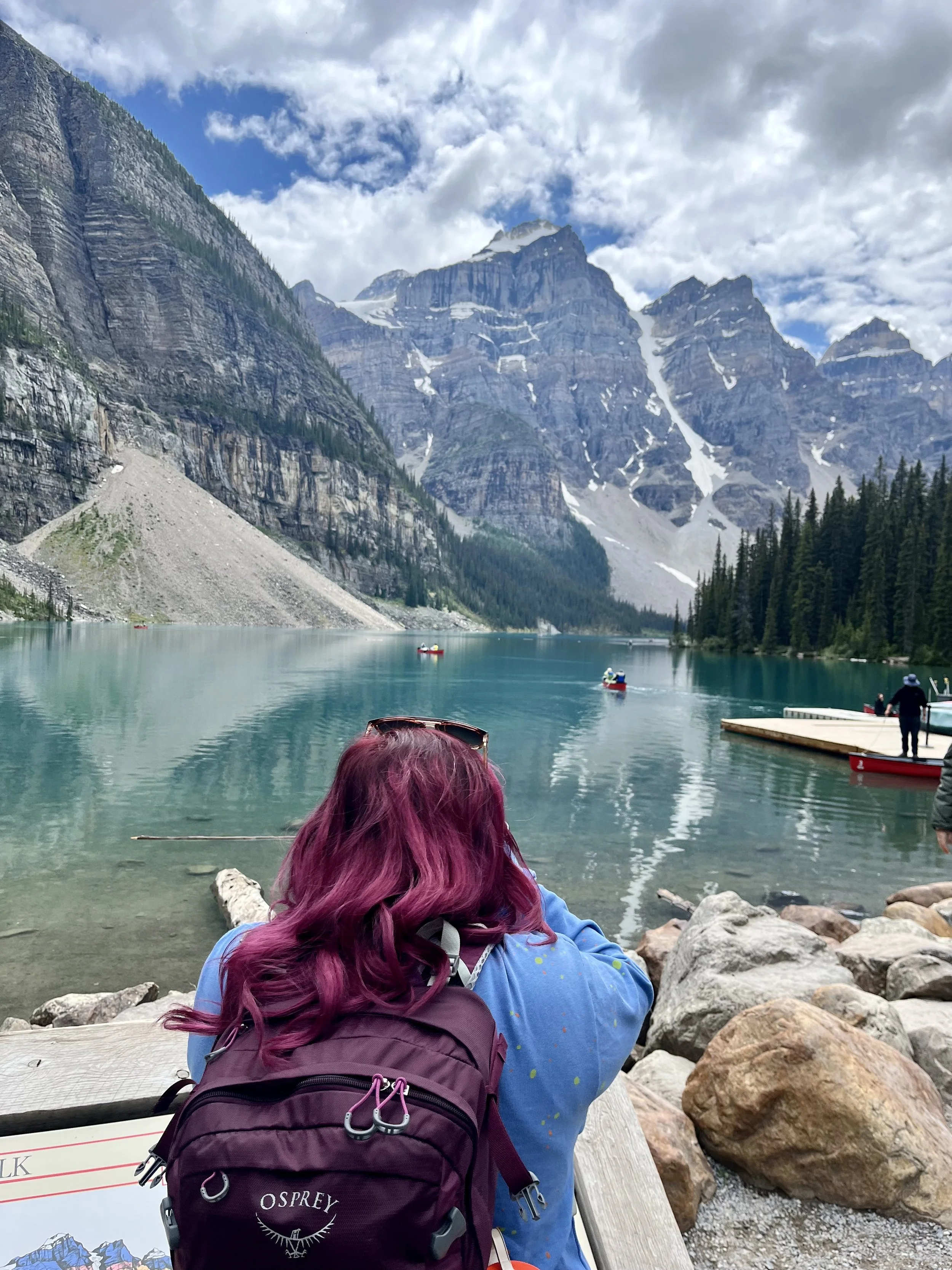 A woman with purple hair and a purple backpack sitting on a wooden bench, overlooking a clear mountain lake with boats, surrounded by tall mountains and pine trees under a partly cloudy sky. Moraine Lake, Banff National Park Canada
