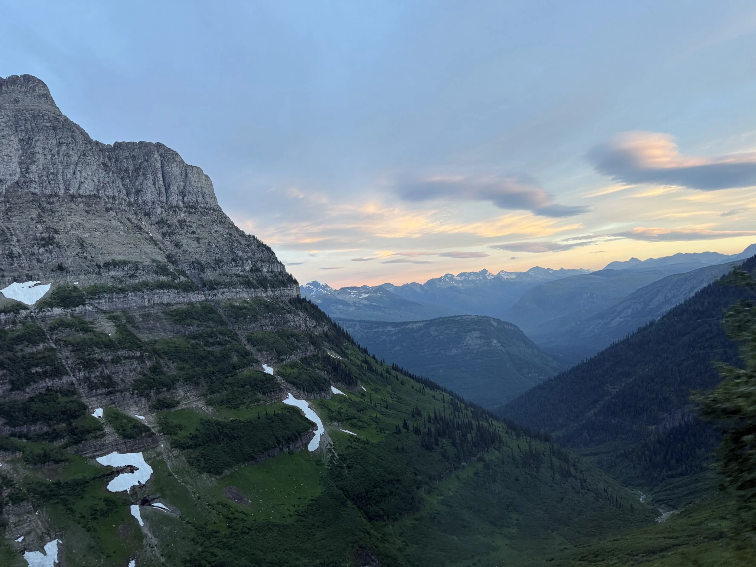 Scenic view of a mountain valley during sunset with rugged mountains, green forested slopes, patches of snow, and a partly cloudy sky.