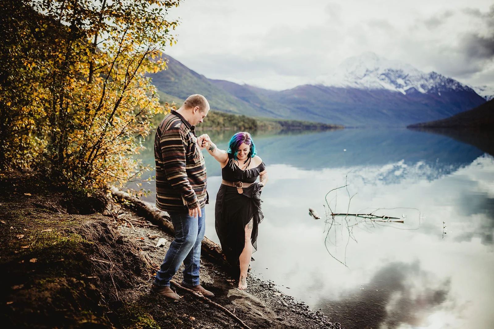 A man lifts a woman by the arm while walking along the edge of a lake surrounded by mountains and trees, with snow-capped peaks in the background. The woman has colorful hair and is wearing a black dress, while the man is dressed casually. The scene 