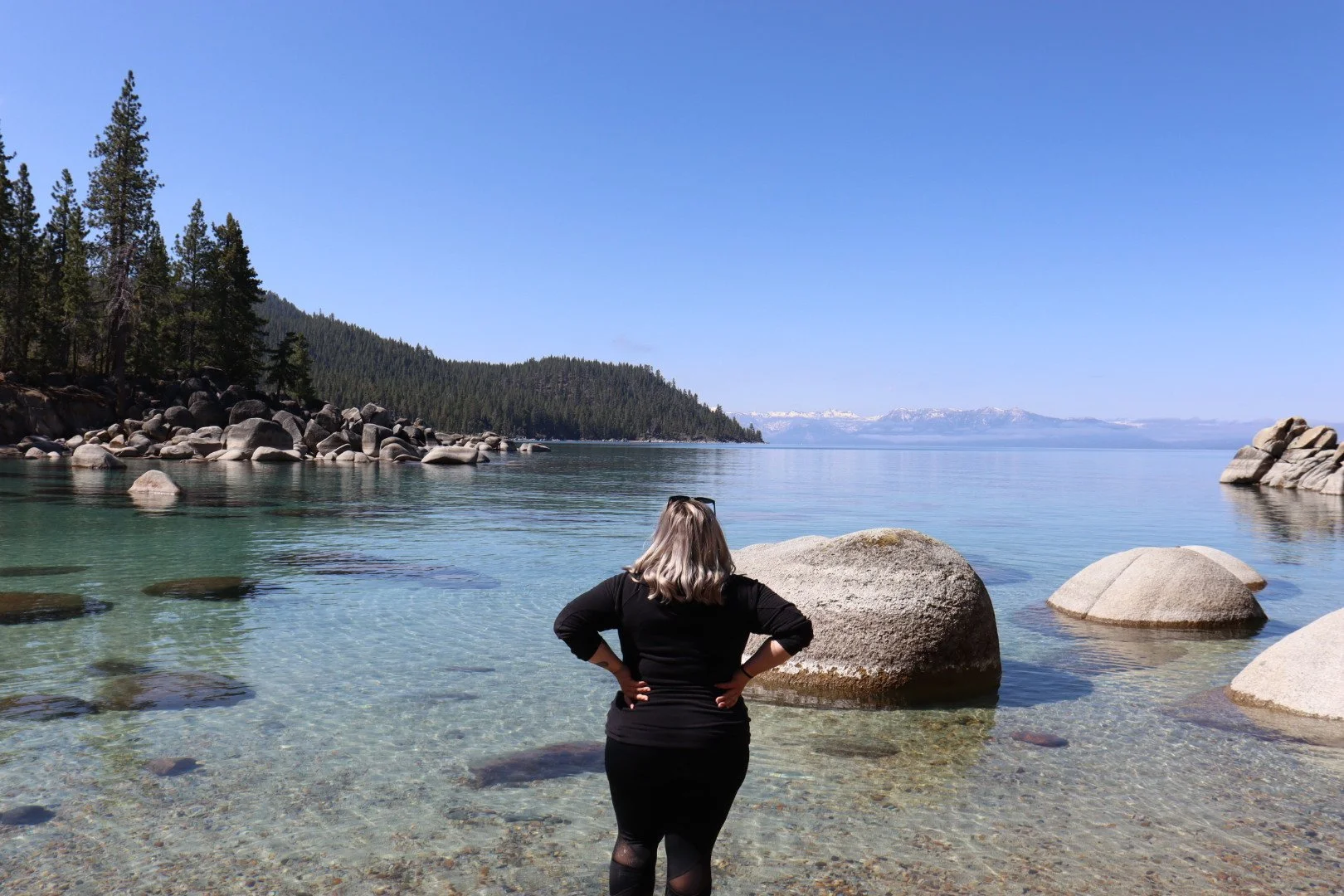 A woman with blonde hair, wearing black clothing, standing on a rocky shoreline, looking out at a calm lake with clear water, surrounded by trees and mountains under a clear blue sky.