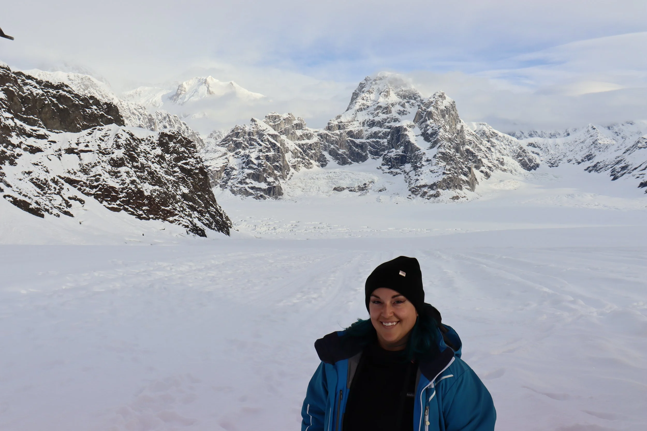 A woman smiling in a blue jacket and black beanie standing in a snowy landscape with snow-covered mountains in the background.