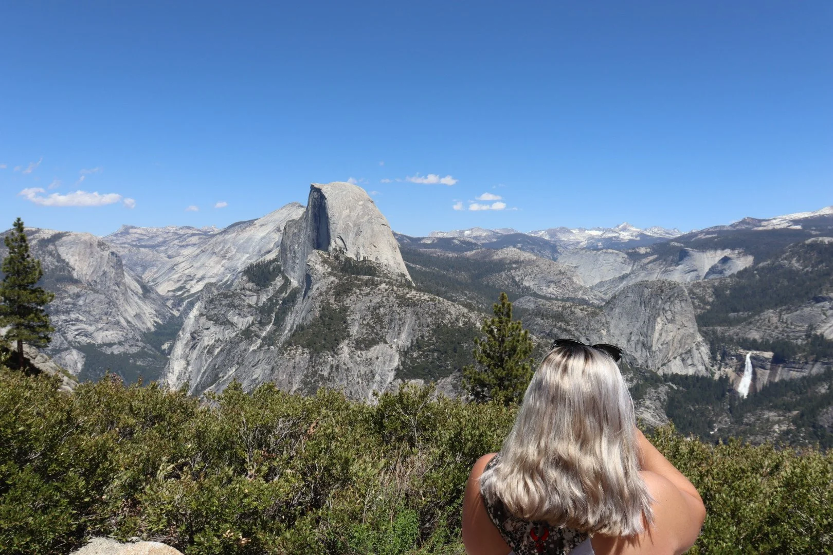 A woman with blonde hair, wearing sunglasses, taking a photo of a mountain landscape with a waterfall in the distance, lush green bushes in the foreground, a clear blue sky, and prominent granite cliffs. Yosemite National Park