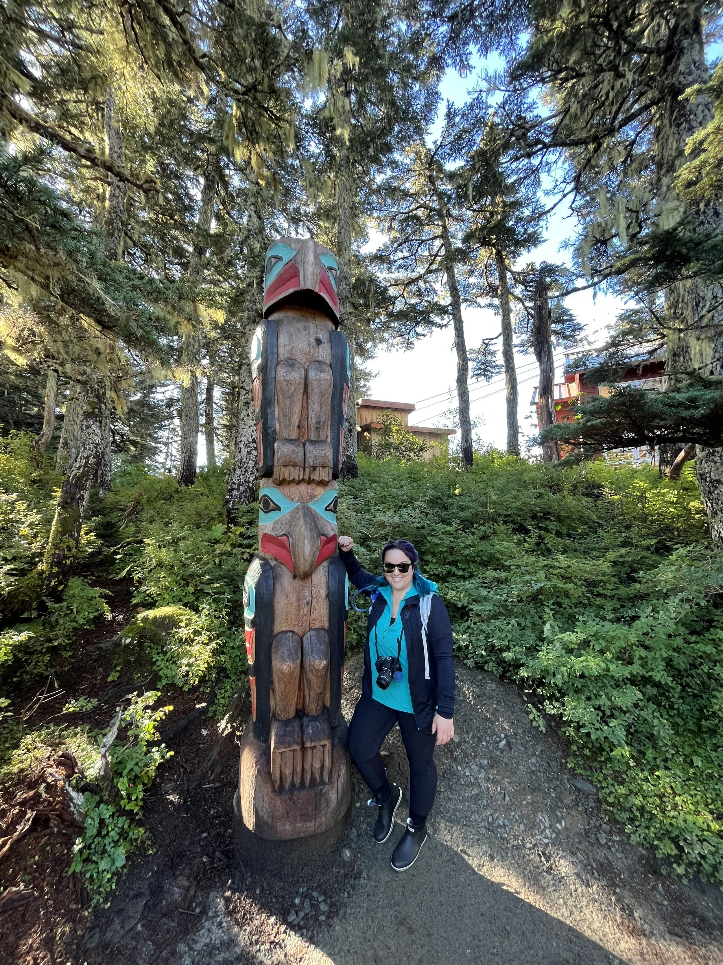 A woman wearing sunglasses, a teal shirt, and a black jacket stands next to a tall totem pole in a forested area, smiling at the camera.