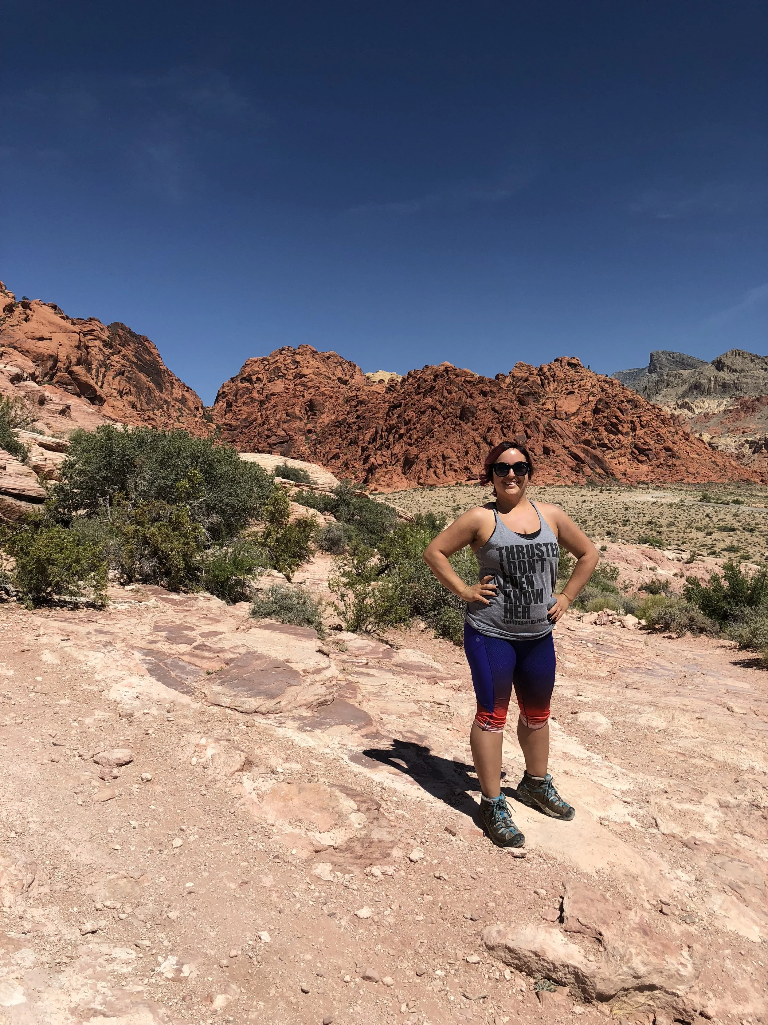 A woman in hiking attire stands on rocky terrain with red rock formations and desert vegetation behind her, under a bright blue sky.