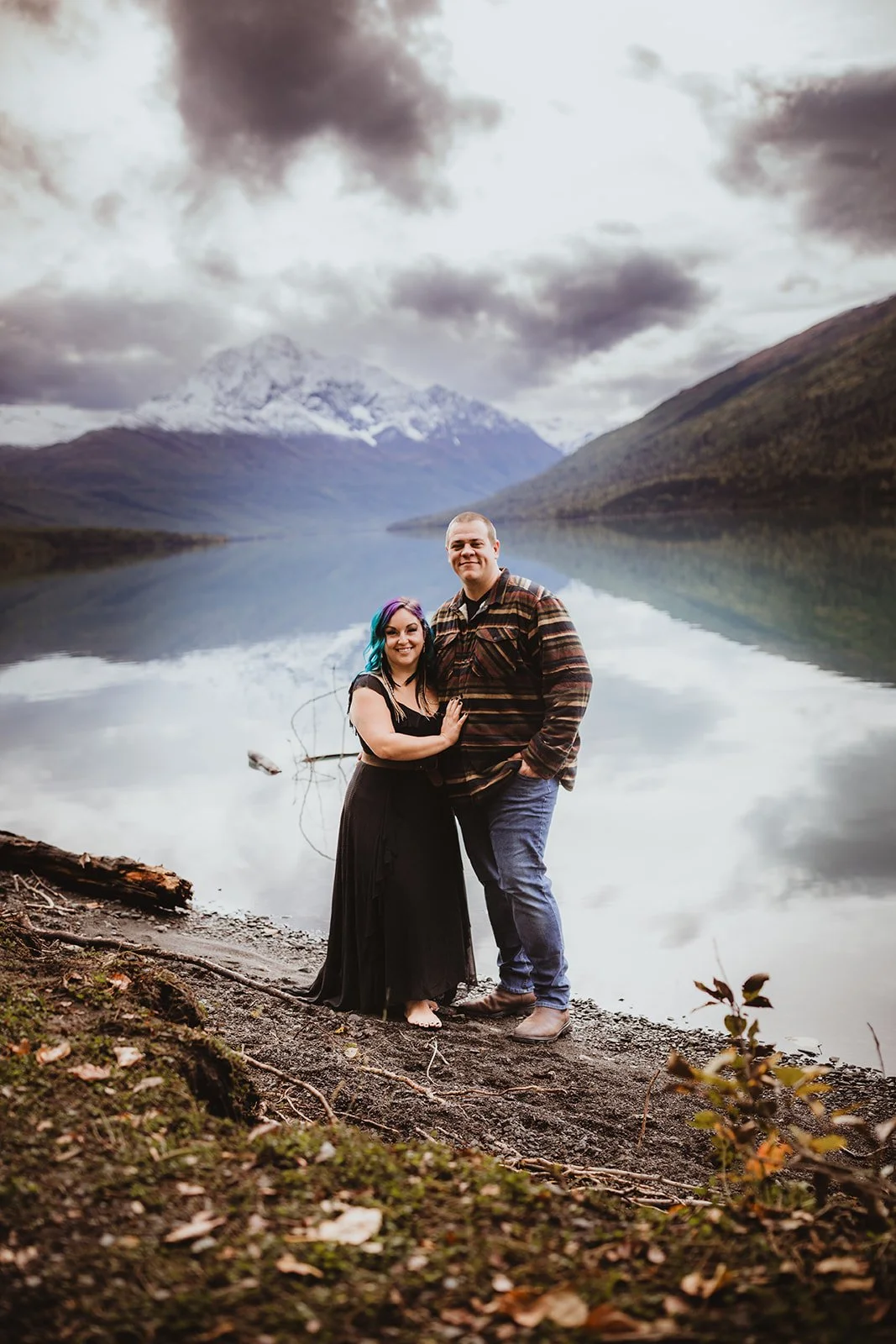 A couple standing by a lake with mountains in the background, smiling at the camera.