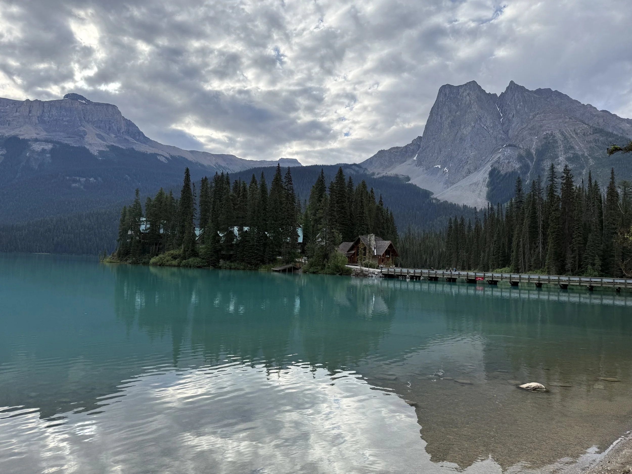 Scenic view of a lake with turquoise waters, a small dock and building, surrounded by evergreen trees, with tall mountains and a cloudy sky in the background.