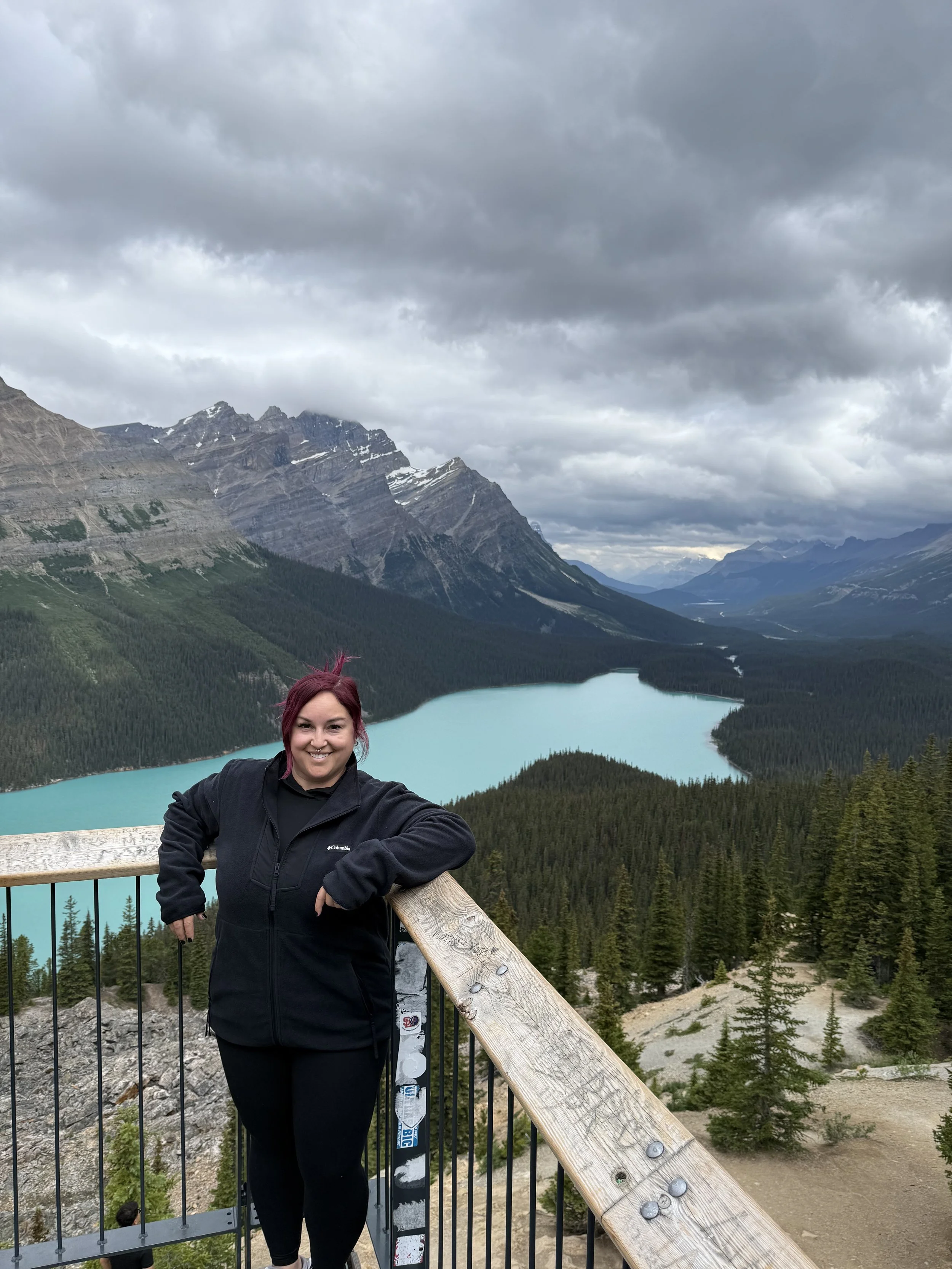 A woman with red hair smiling and wearing black outdoor clothing, leaning on a wooden railing at a scenic mountain overlook with a turquoise lake, dense forest, and rugged mountains under a cloudy sky in the background. Peyto Lake Canada