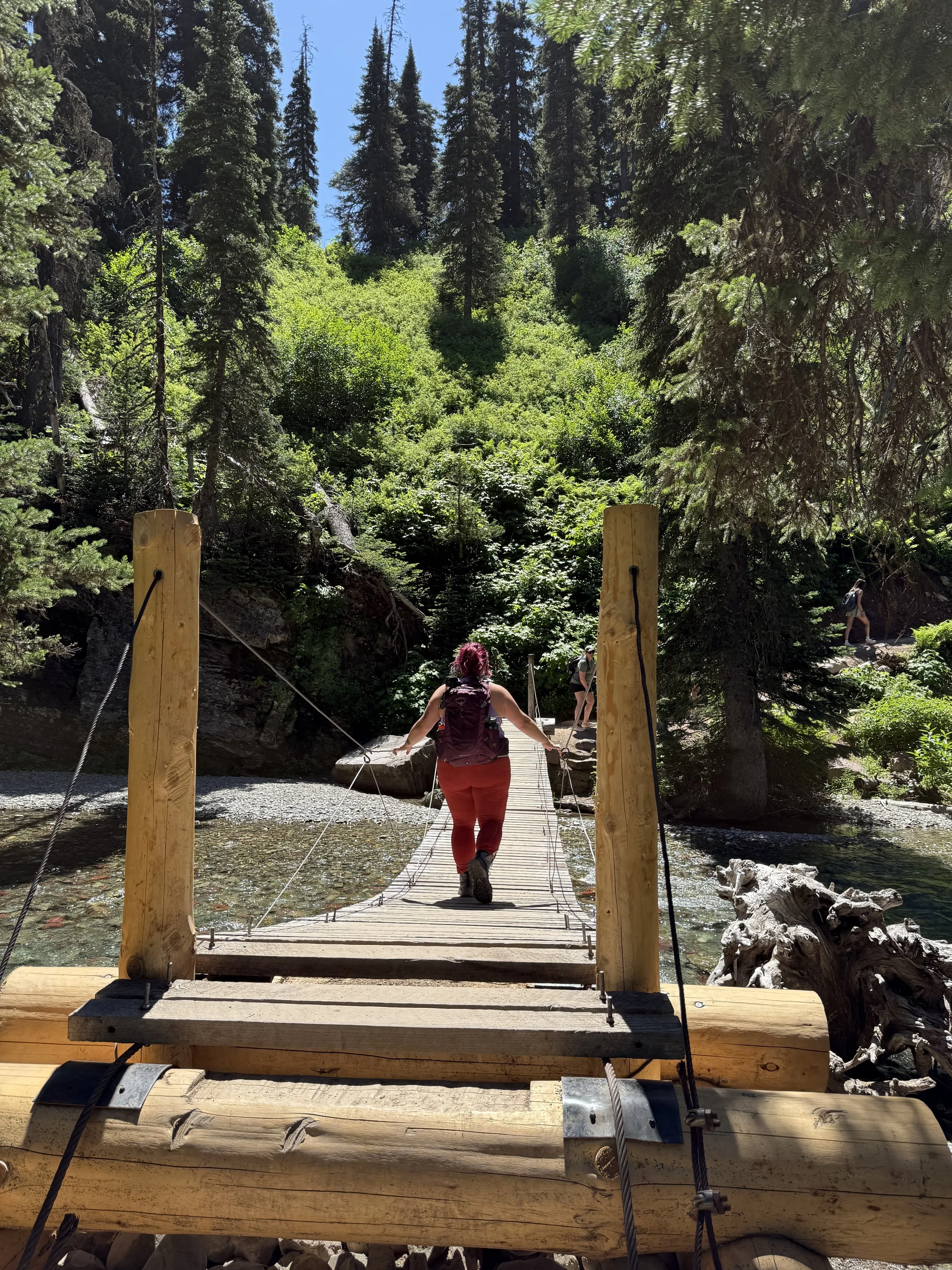 A person crossing a wooden suspension bridge over a river in a forest surrounded by tall trees and lush green foliage.