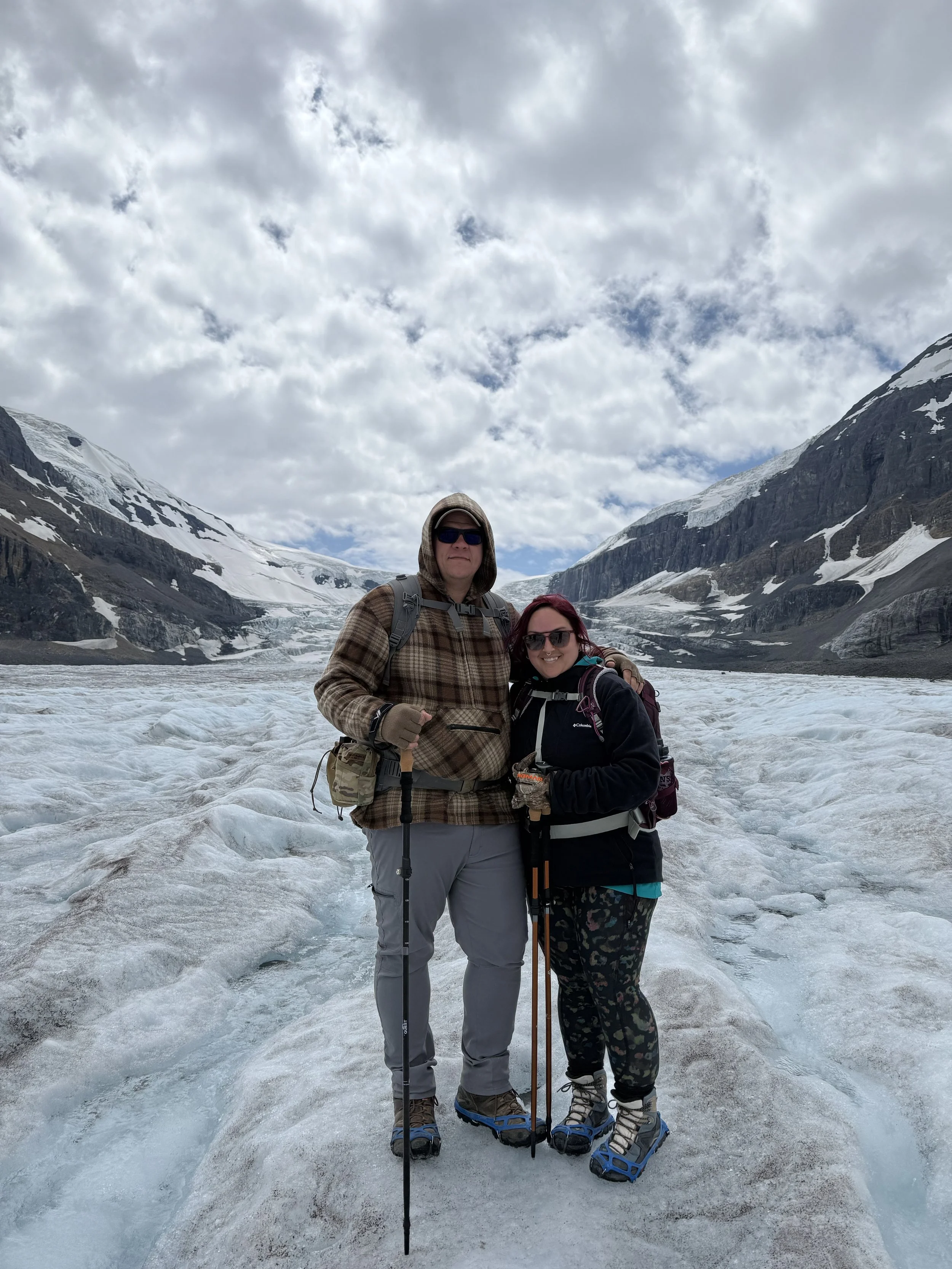 Two people standing on a glacier in a mountainous area with snow-covered peaks, wearing hiking gear and sunglasses. Athabasca Glacier, Canada