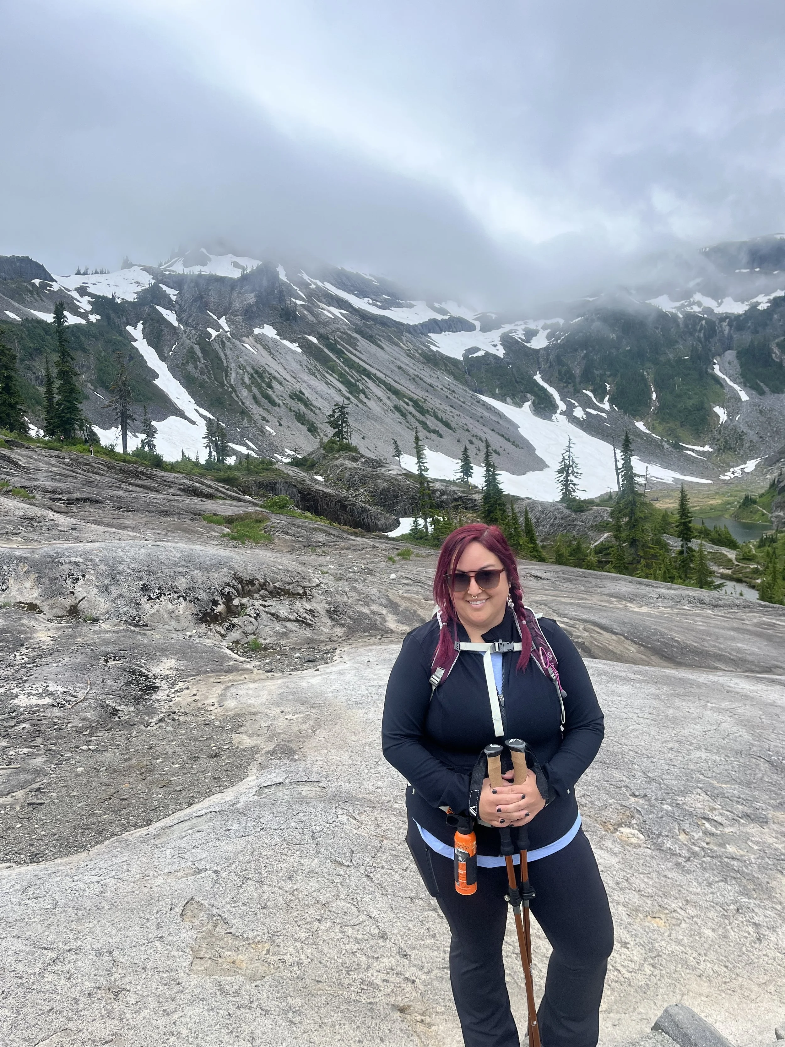 Woman with sunglasses and purple hair hiking on rocky terrain in a mountainous area with snow patches and tall pine trees, under cloudy sky.
