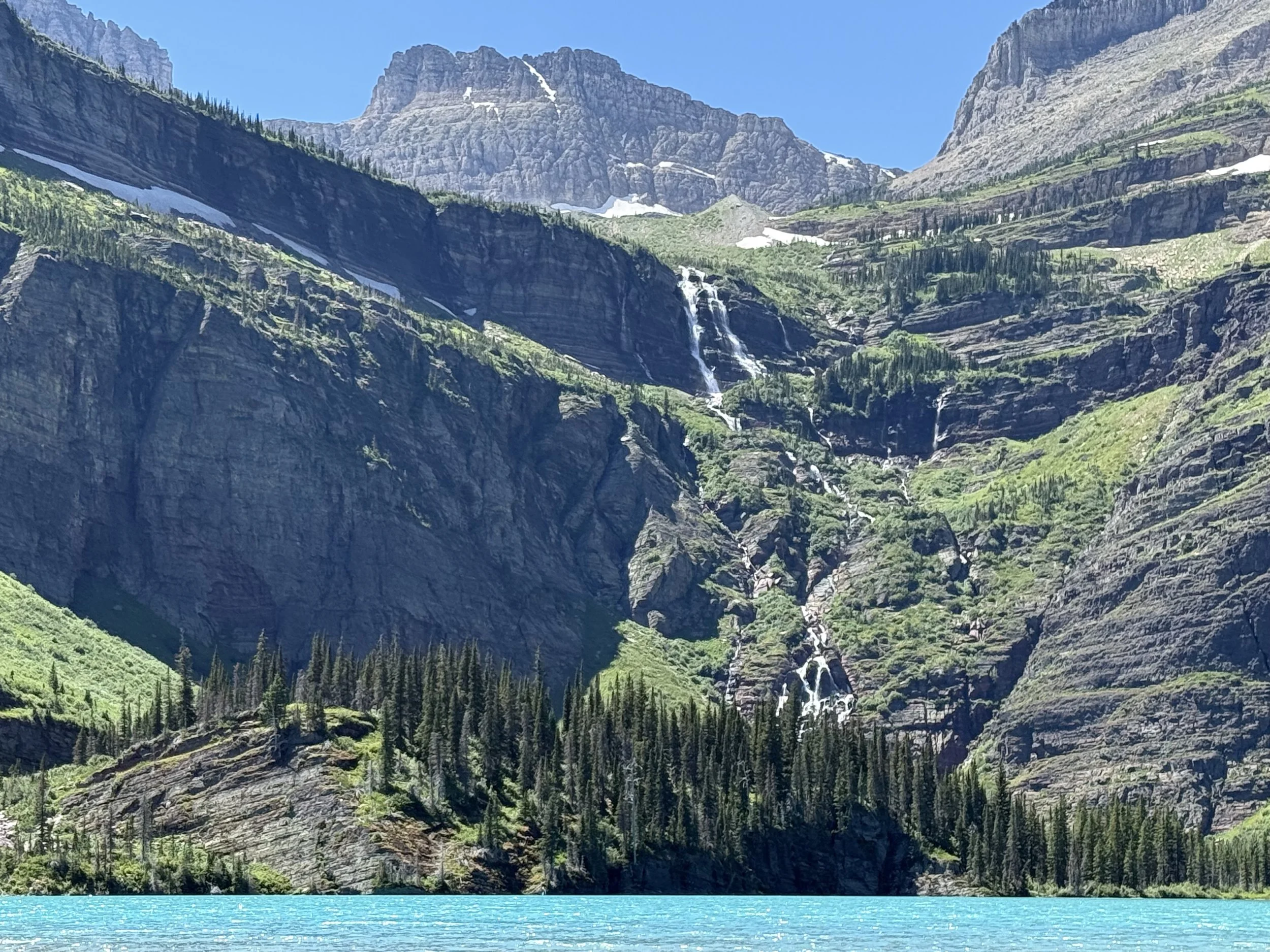 A mountain landscape featuring a large body of turquoise water in the foreground, densely forested lower slopes, rocky cliffs, and a cascading waterfall on lush green mountains with patches of snow at the peaks under a clear blue sky.