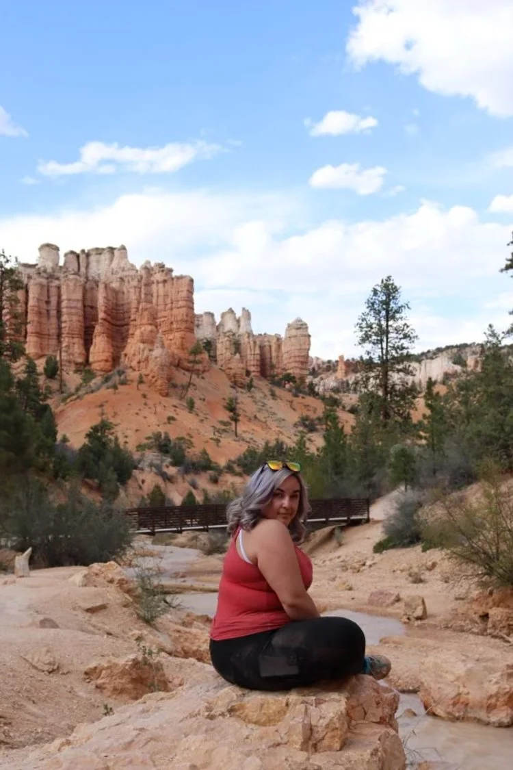 A woman with gray hair sitting on a rock in a desert landscape. Behind her are orange rock formations, a small creek, trees, and a wooden bridge. The sky is partly cloudy. Bryce Canyon National Park