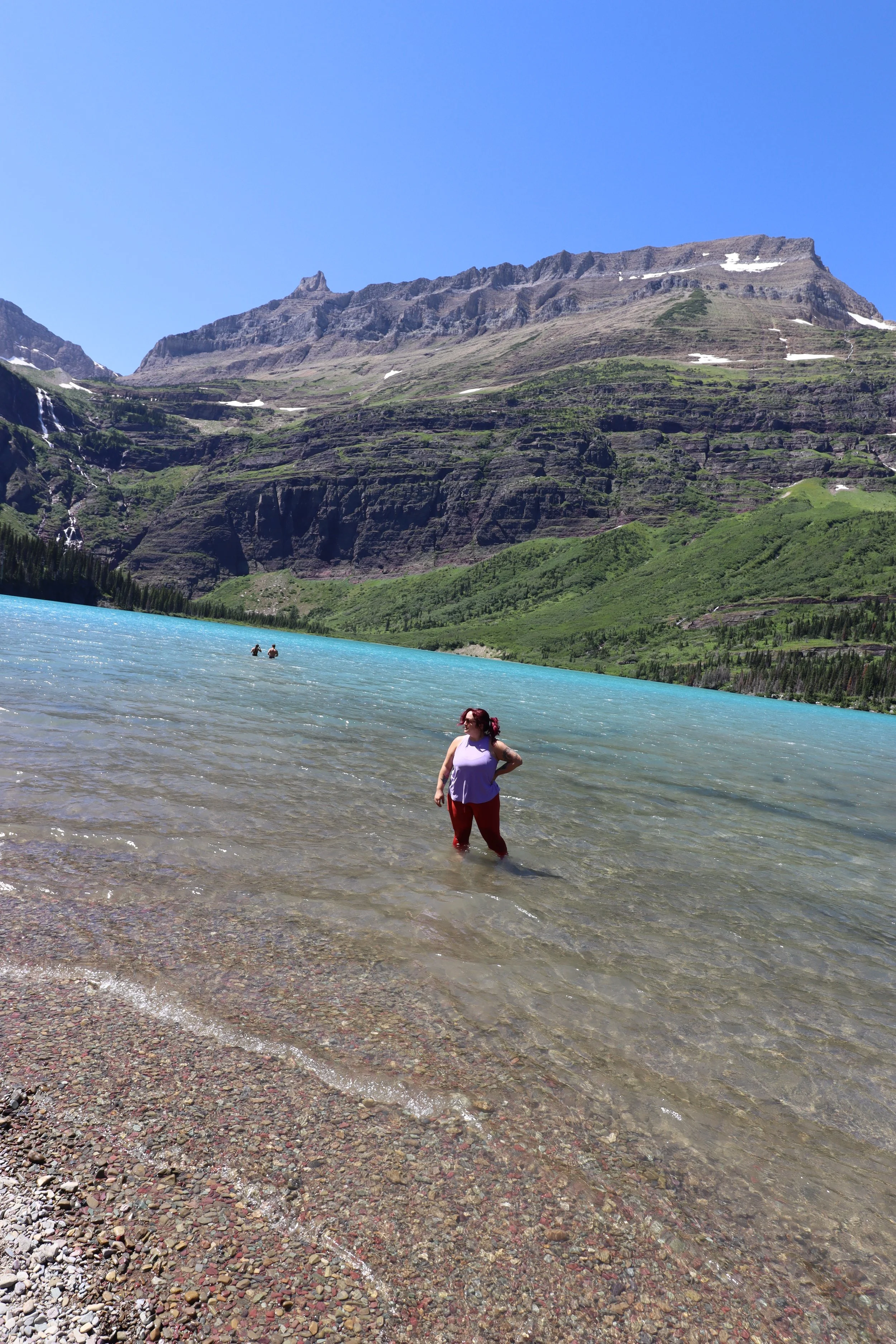 A person in a purple top and red pants standing in a clear lake with a mountainous landscape in the background, under a bright blue sky.