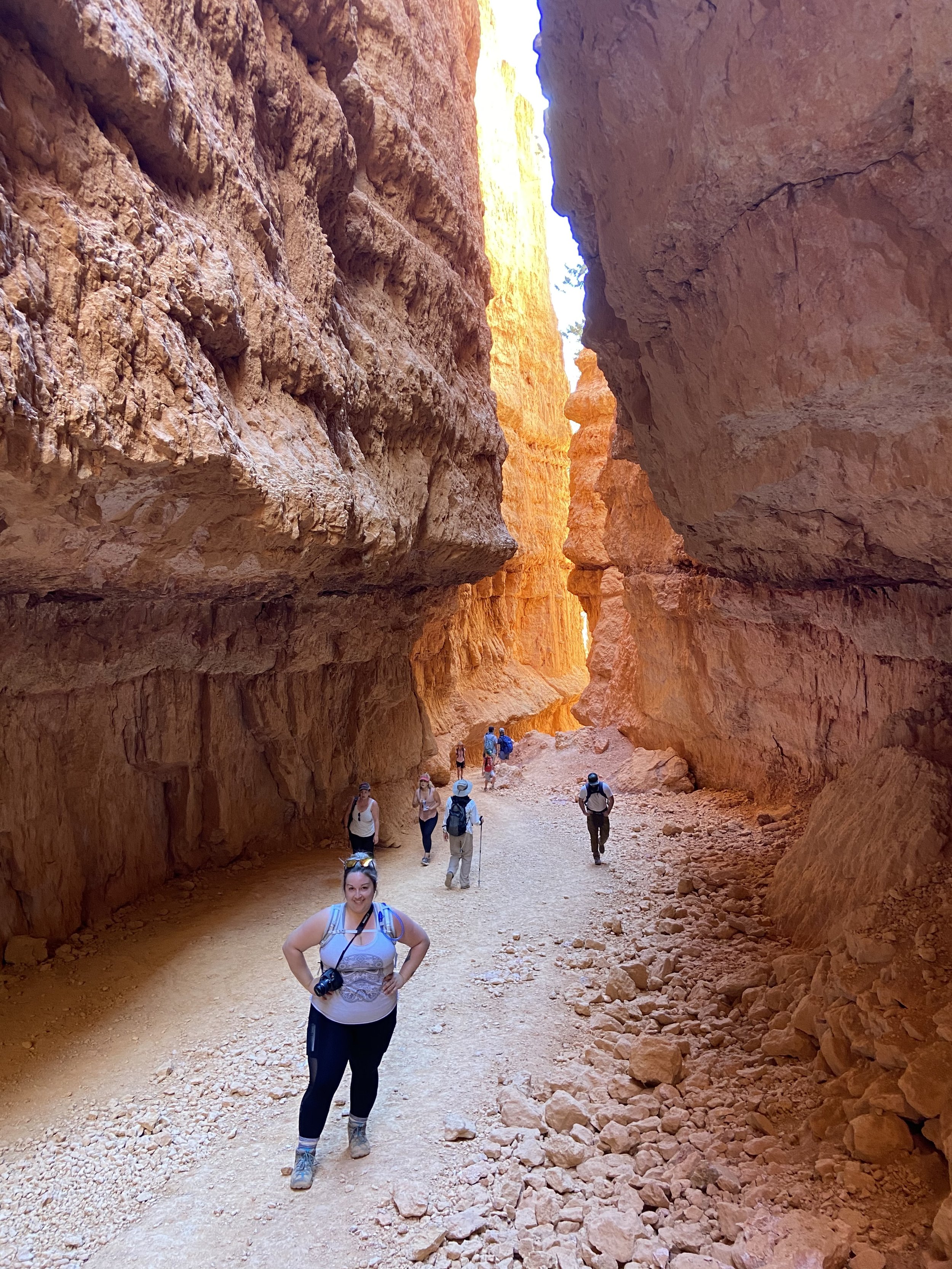Group of hikers walking through a canyon with tall, orange and red rock walls, some sunlight shining on the rocks.