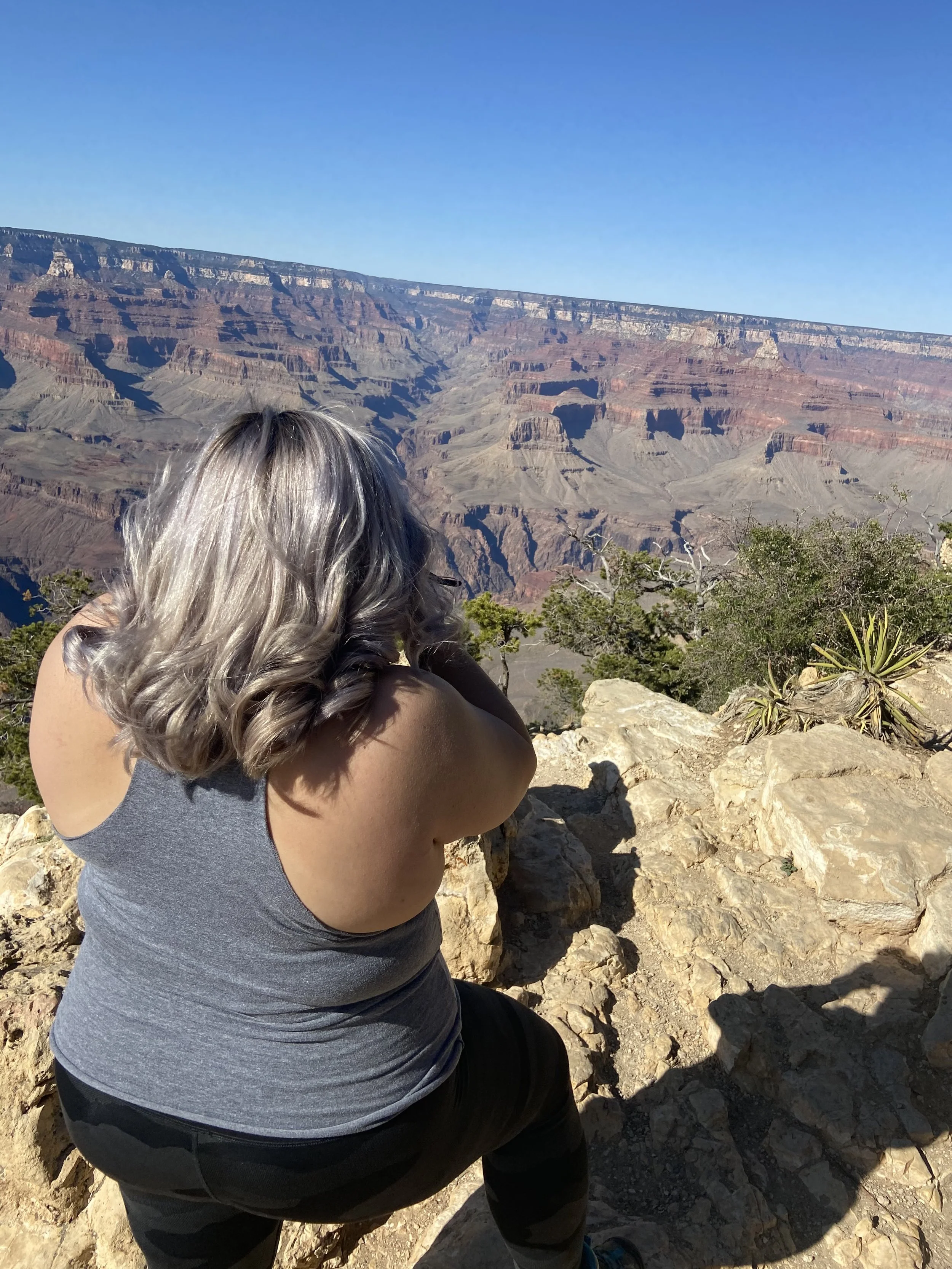 A woman with gray hair taking a photo of the Grand Canyon with her phone from a rocky viewpoint.
