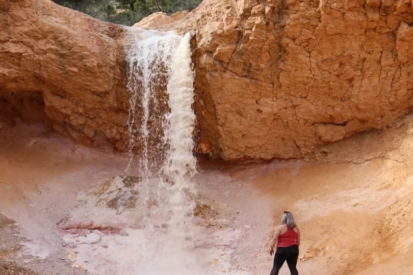 A woman standing in front of a small waterfall flowing down a reddish, rocky canyon.