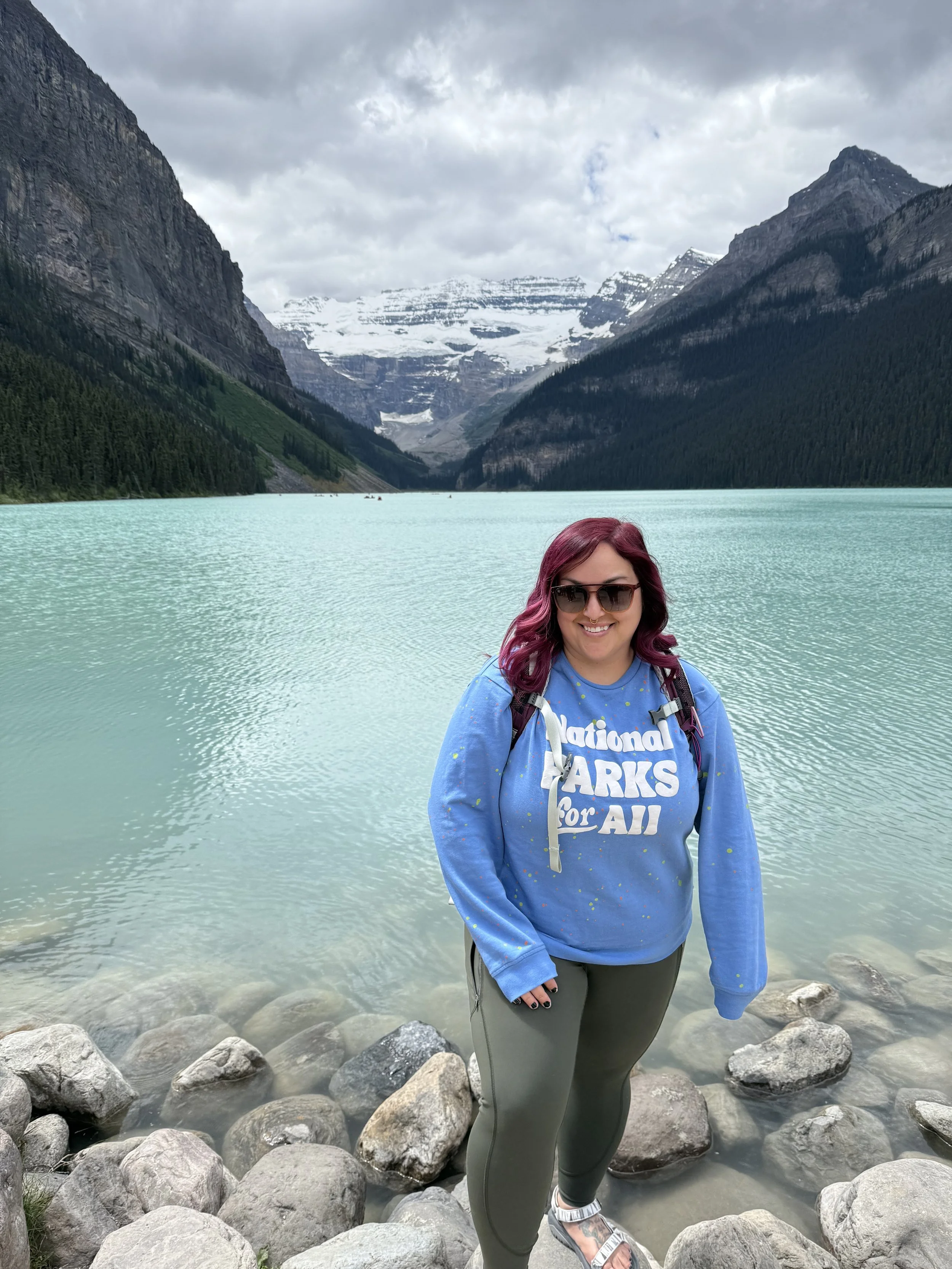 Woman with purple hair and sunglasses standing on rocks at the edge of a turquoise lake, surrounded by mountains with snow caps and a cloudy sky.