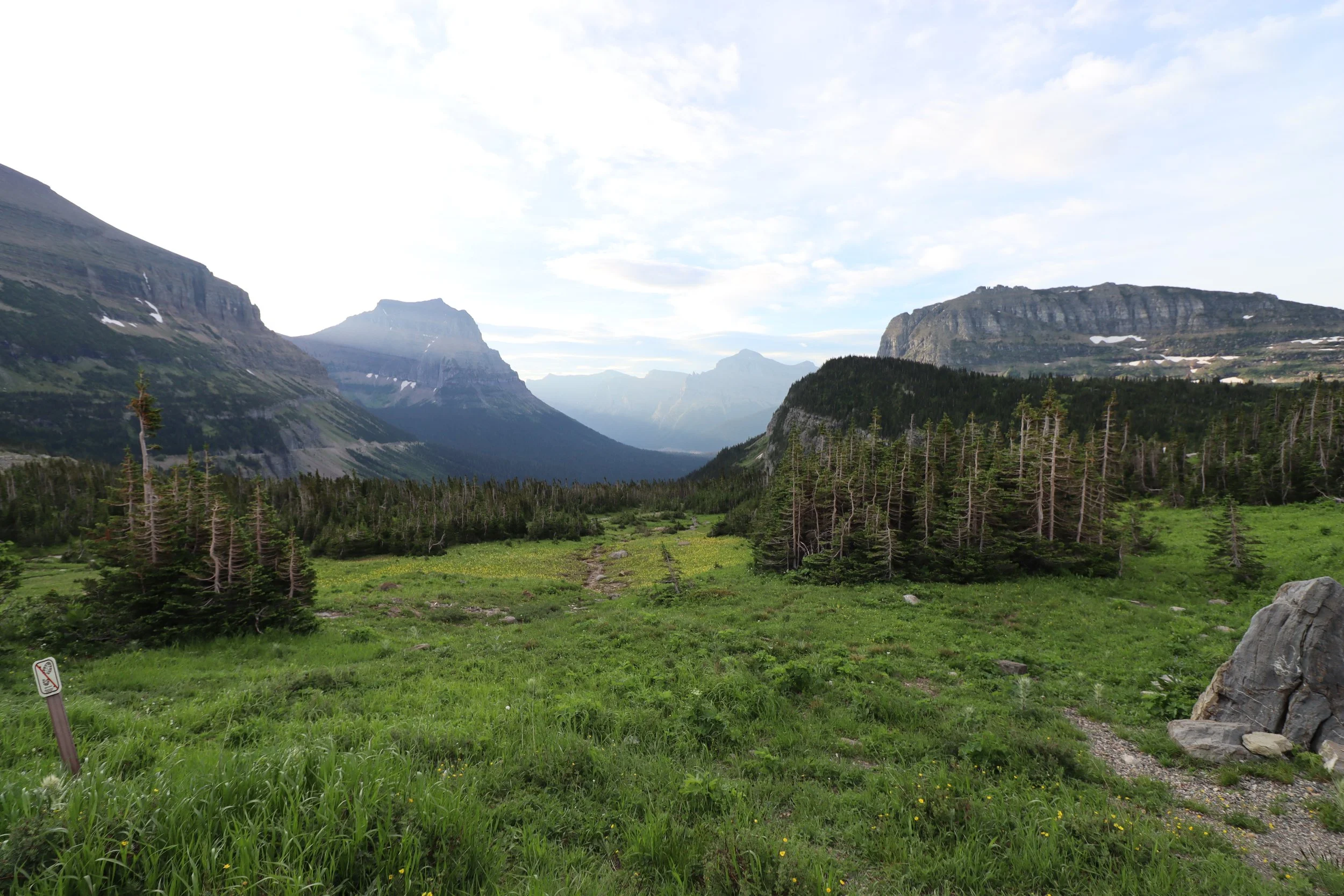 A lush green valley surrounded by tall mountains with patches of snow under a partly cloudy sky.