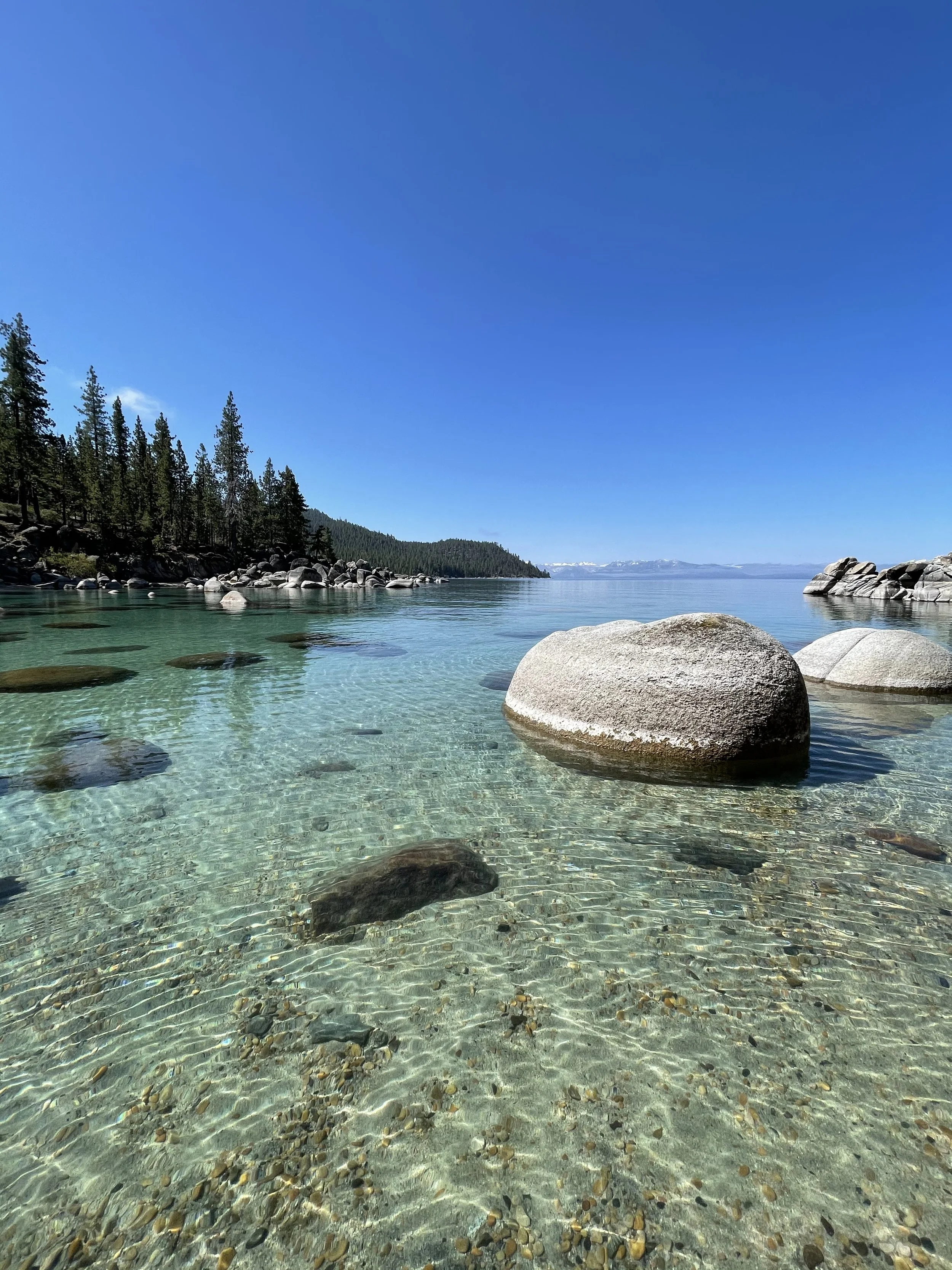 Clear lake with large rocks in the foreground, surrounded by trees and mountains in the distance, under a bright blue sky. Lake Tahoe, CA