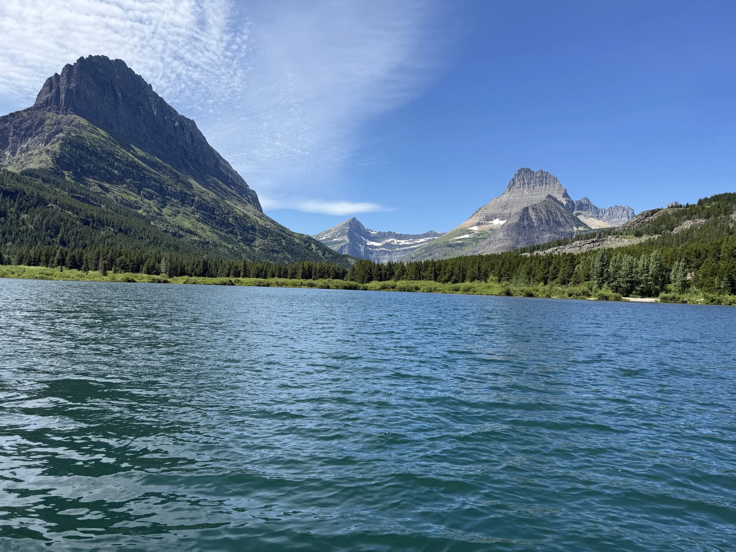 A scenic view of a large lake with turquoise water, surrounded by green forest and mountain peaks under a blue sky with some clouds.