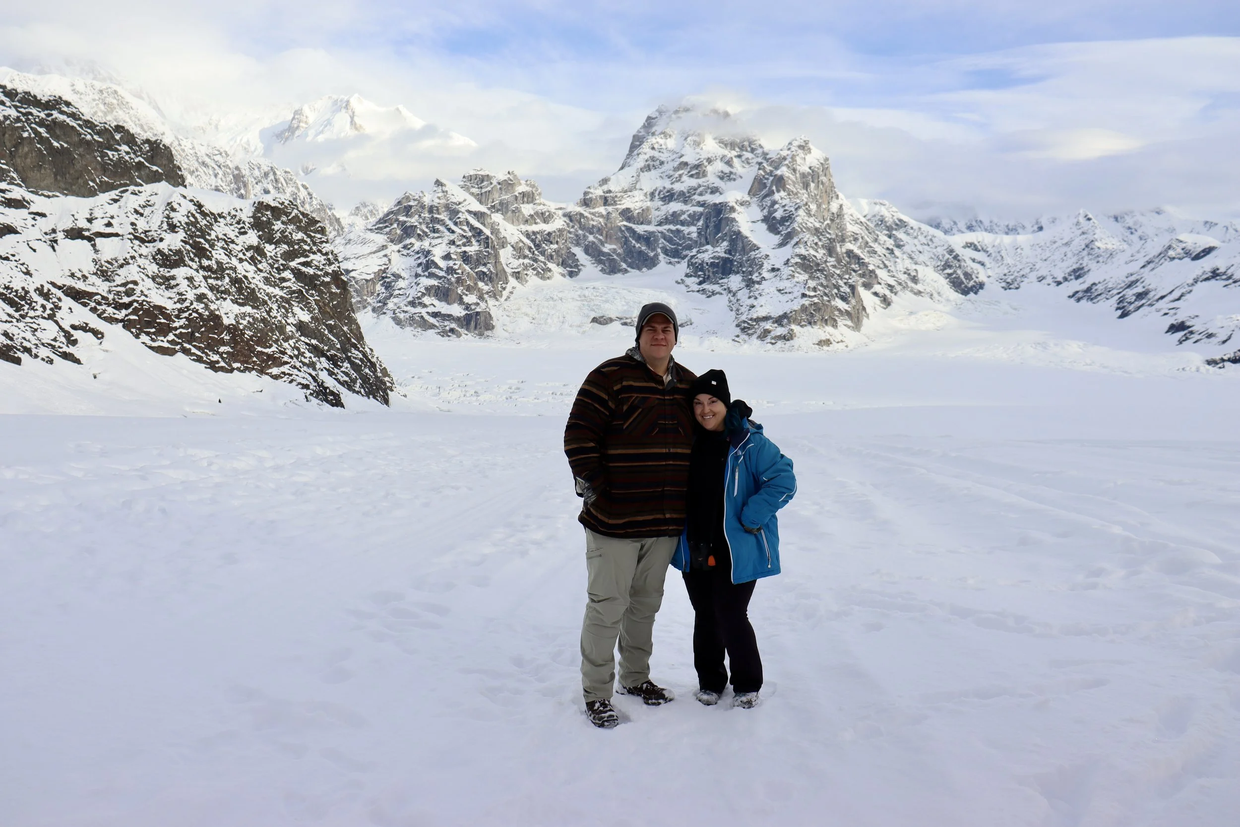 Two people standing in snow with mountains and glaciers in the background