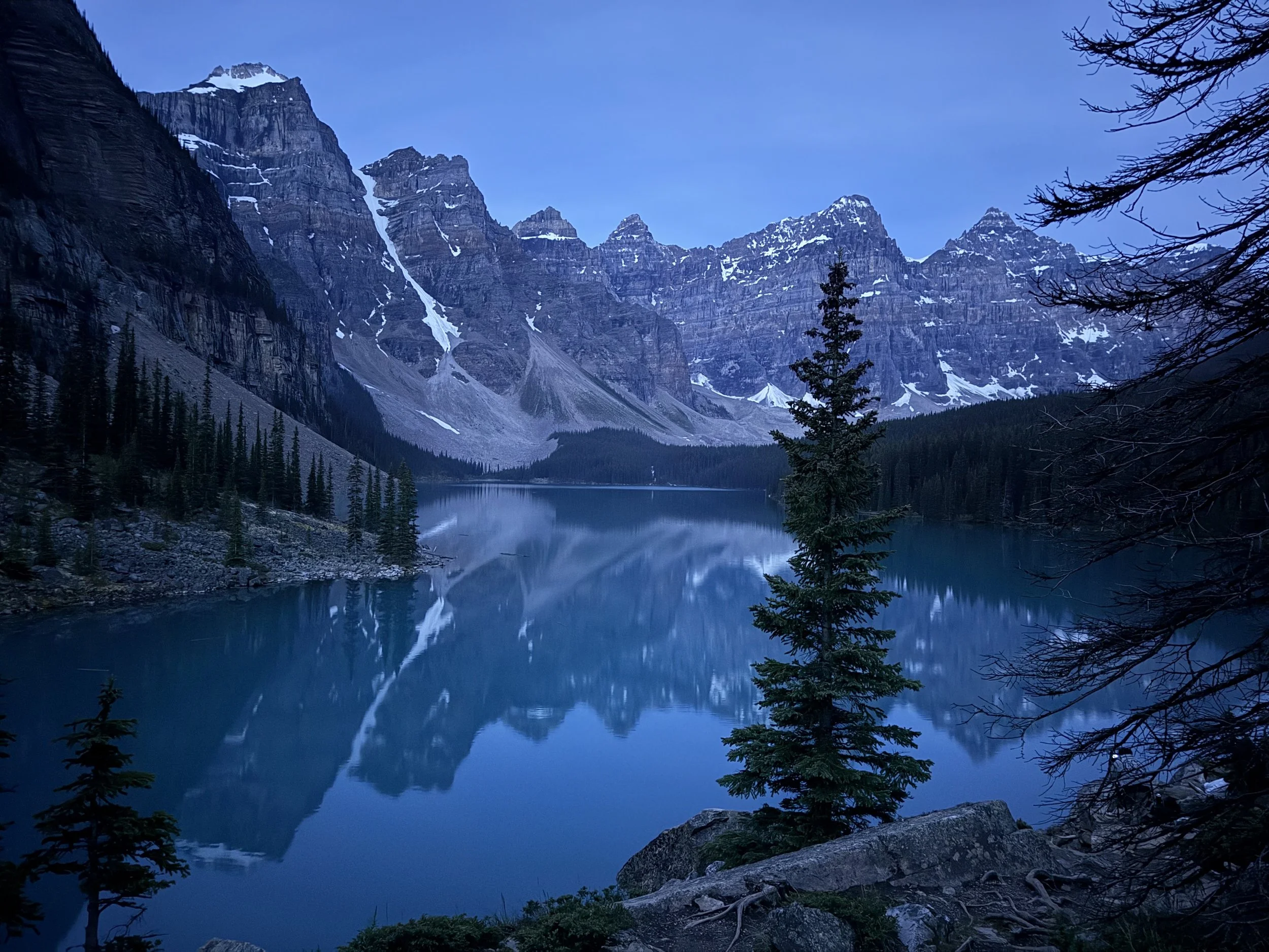 Mountain range with snow patches, reflecting in a calm lake with pine trees along the shoreline in a forested area. Moraine Lake, Banff National Park Canada