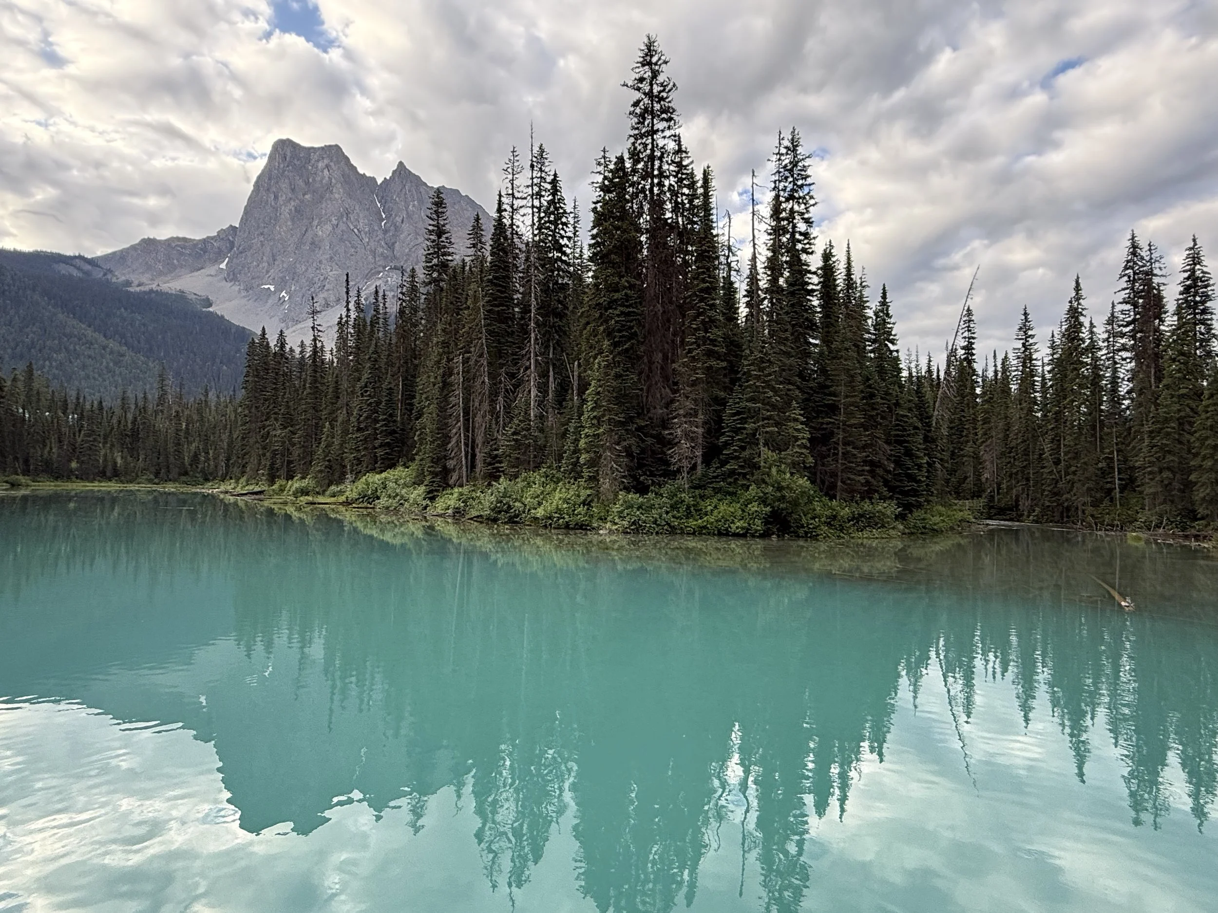 A serene mountain lake surrounded by evergreen trees and topped with a rocky mountain peak under a partly cloudy sky.