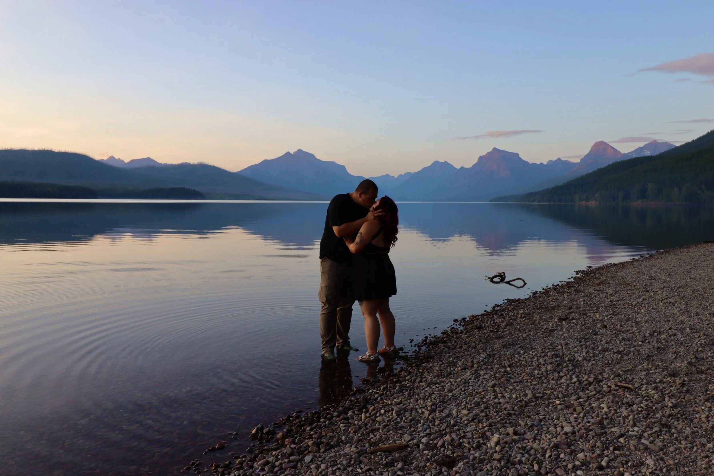 A couple standing in knee-deep water by a rocky shore, sharing a kiss during sunset with mountain ranges and a calm lake in the background.