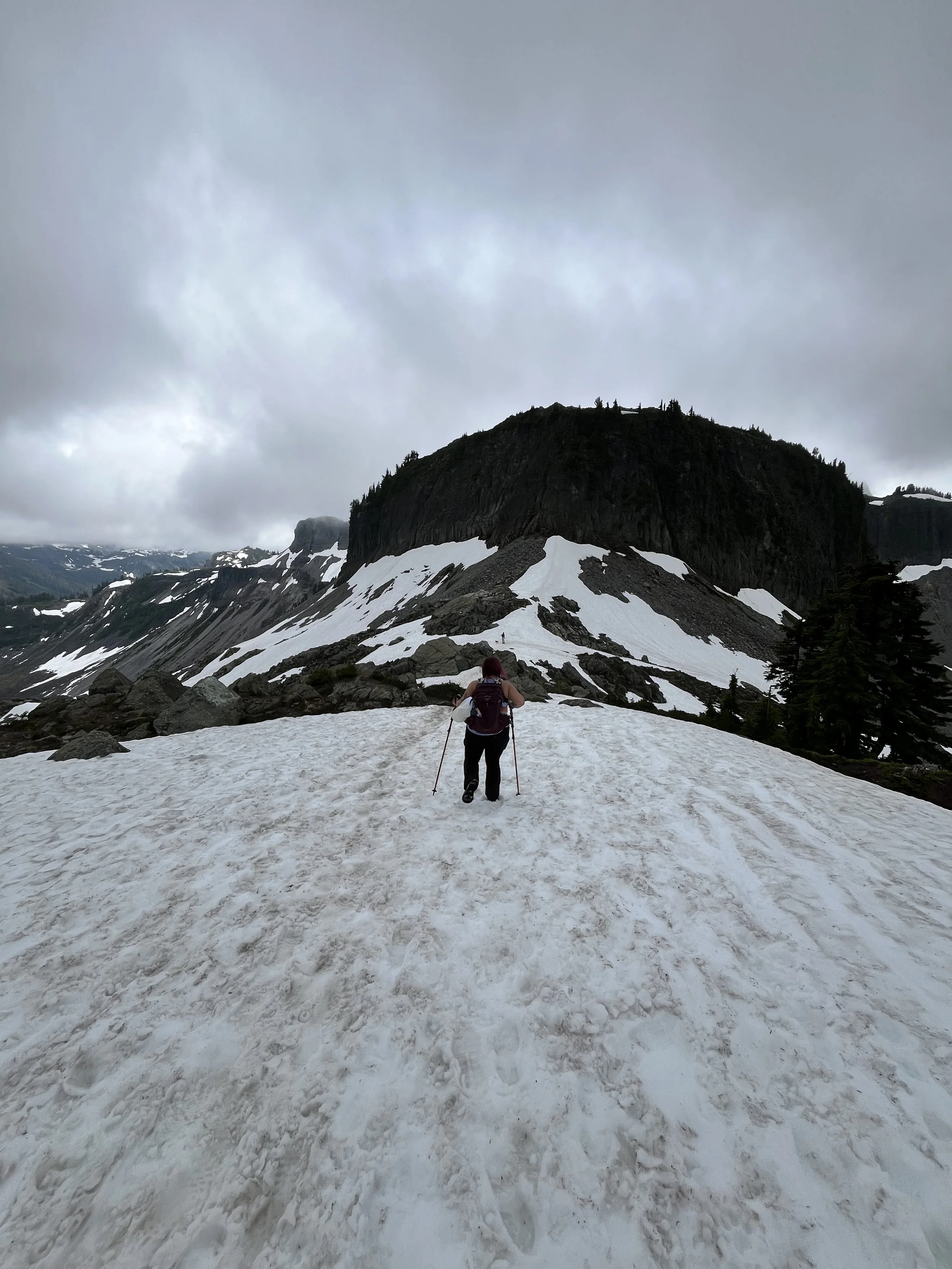 A person hiking on snow-covered terrain with a mountain in the background, and cloudy skies overhead.