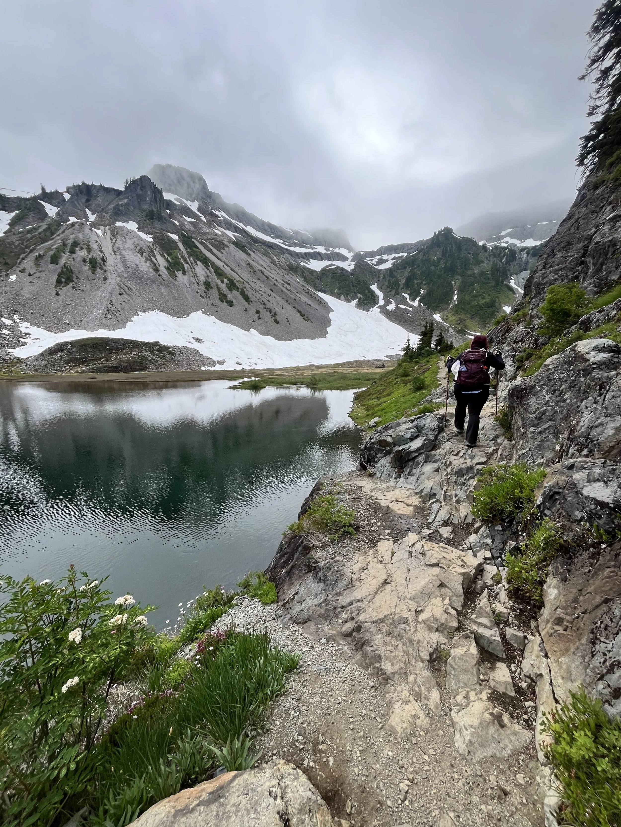 A hiker walking along a rocky trail next to a mountain lake, with snow-capped mountains and cloudy skies in the background.