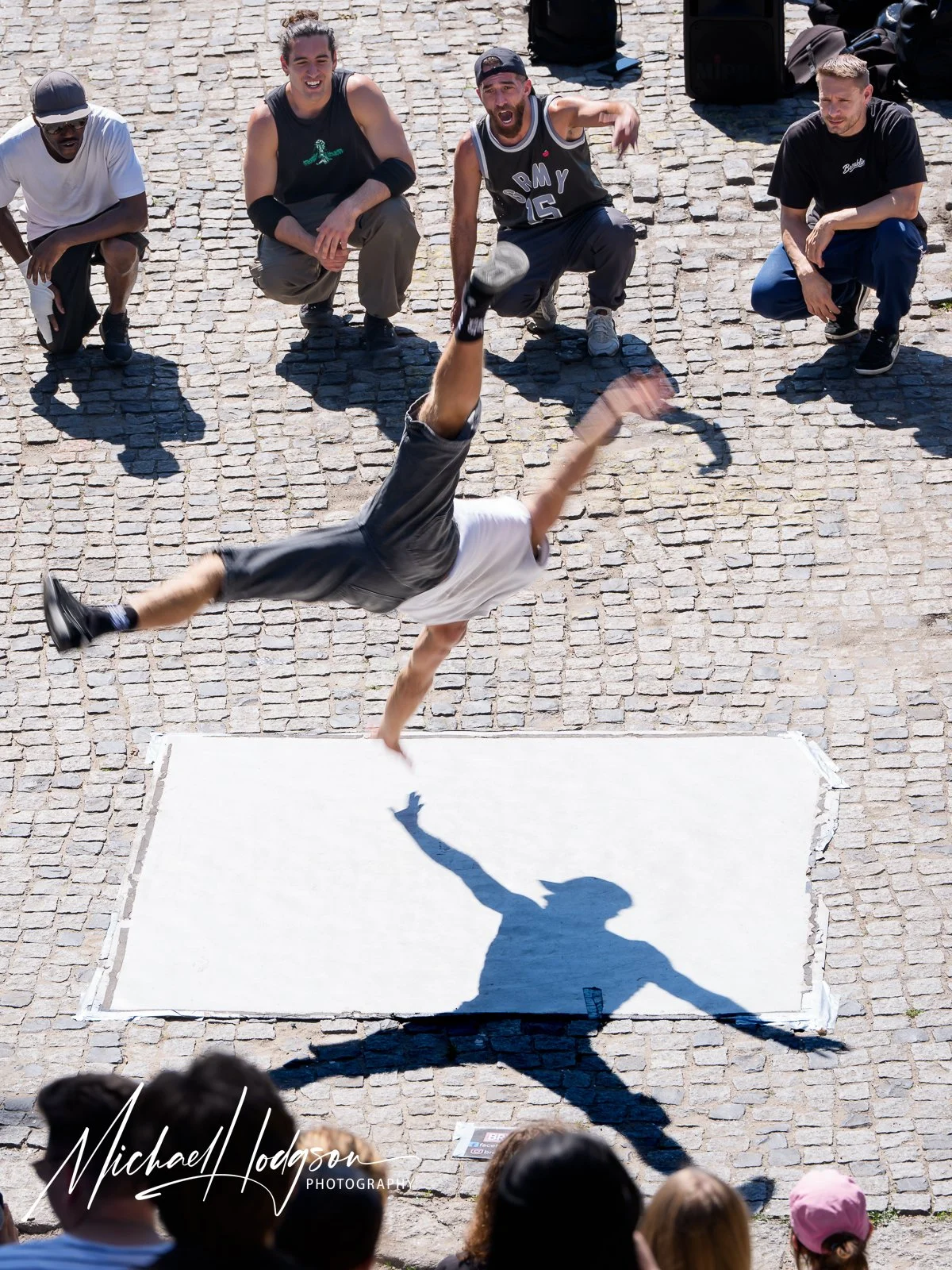 A breakdancer launches into an acrobatic move during a street performance at Mauerpark in Berlin, Germany, while his sharply defined shadow appears to dance across the pavement below.