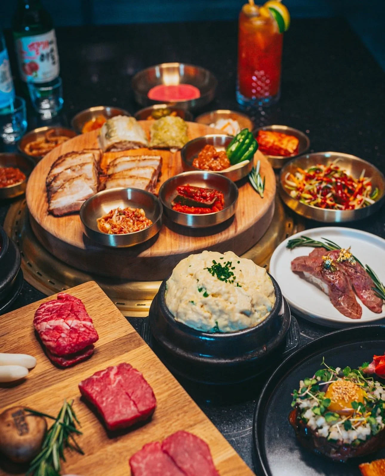 Korean barbecue spread with various raw meats, side dishes including kimchi, potato salad, and salads, alongside drinks on a black table.