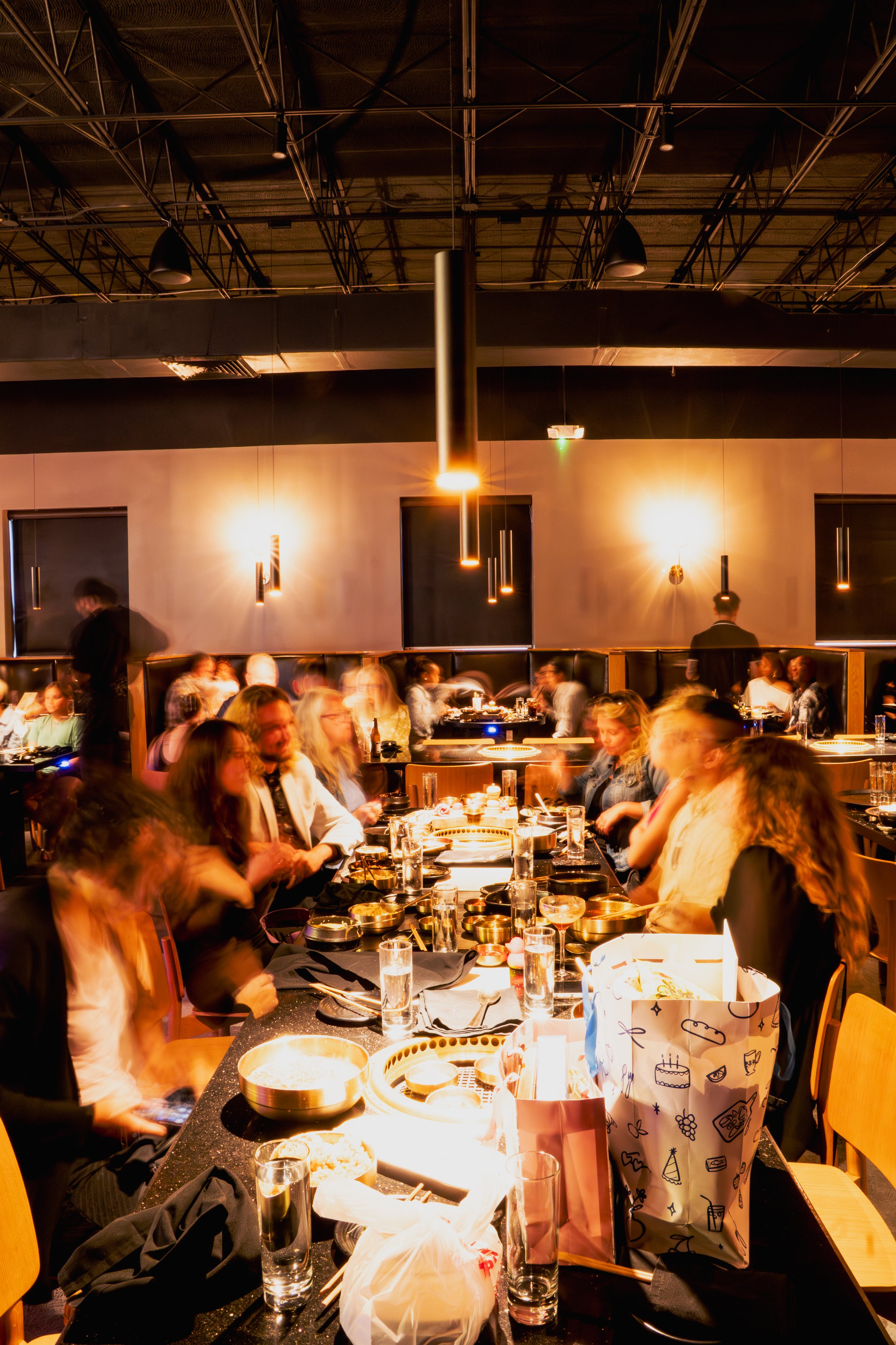 A busy restaurant with many people dining at tables, some engaging with their phones. The decor features black walls with hanging lights, an exposed ceiling, and a vibrant, warm lighting throughout the space.